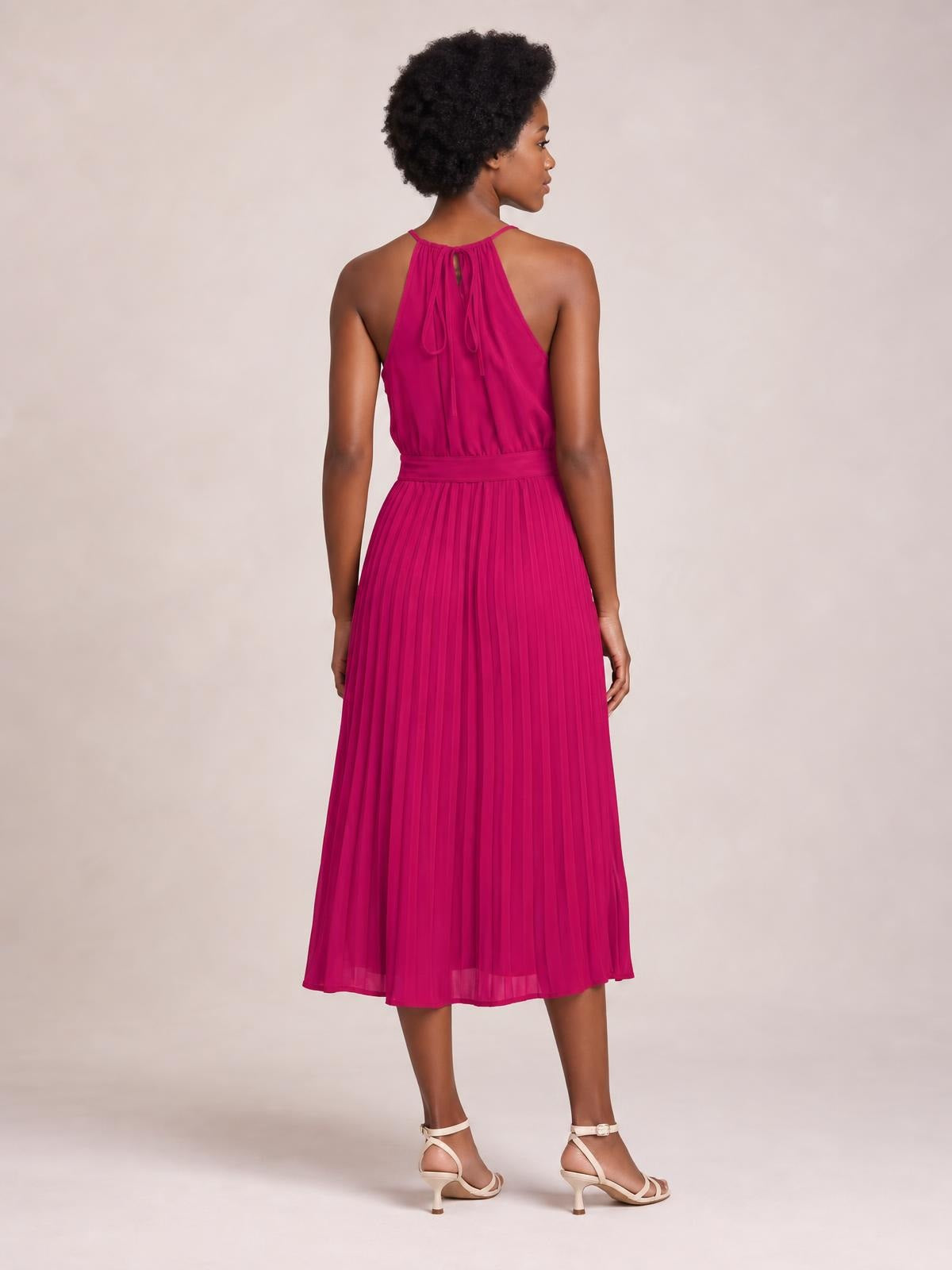 A model poses from the back in a vivid magenta pleated midi dress with thin tie straps, a defined waist, and a softly flared pleated skirt.