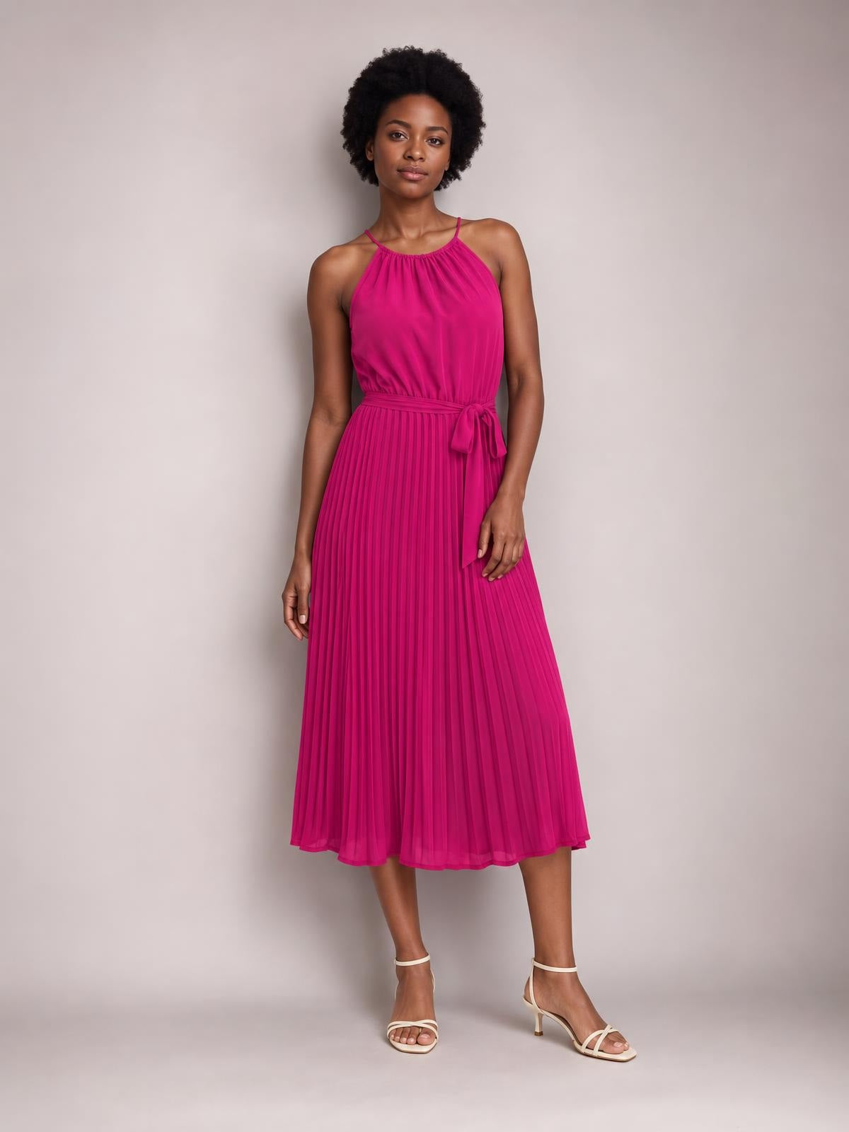 Model poses in a bright fuchsia pleated spaghetti strap midi dress with a tied waist, gathered neckline, and softly flared silhouette.