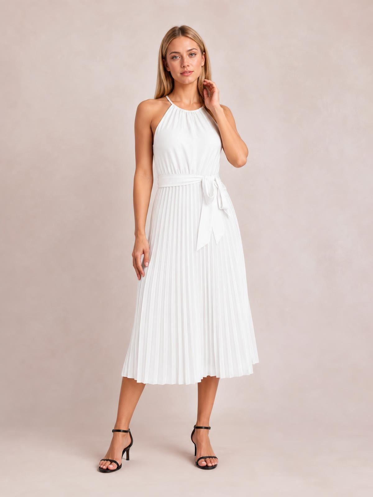 Model poses in a white pleated spaghetti strap midi dress with a halter-style neckline, defined waist, tied sash, and flowing A-line silhouette.