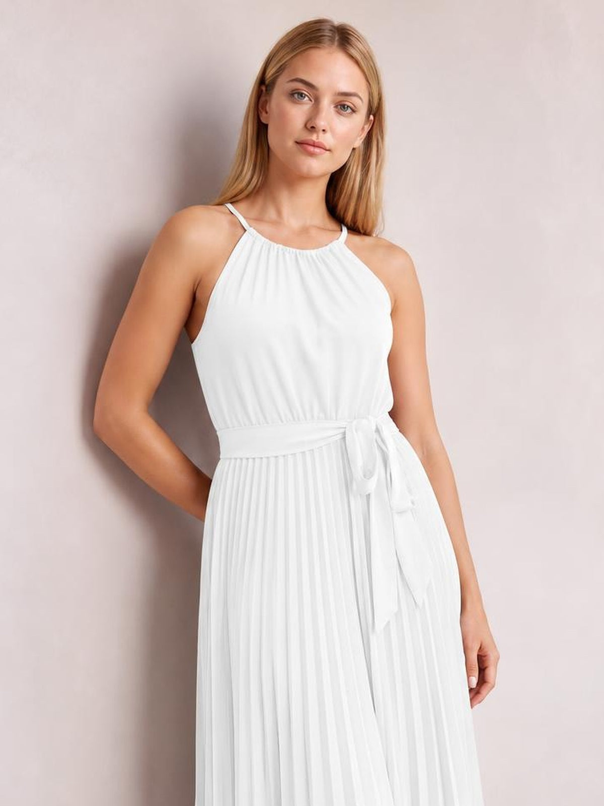 Model wearing a white pleated spaghetti strap midi dress with a defined waist, tied sash, flowing silhouette, and one hand resting at her hip.