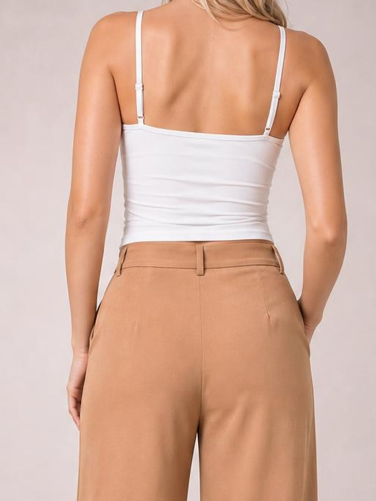 A model poses from behind in camel pleated wide-leg pants with a tailored waistband, belt loops, side pockets, and a relaxed silhouette.
