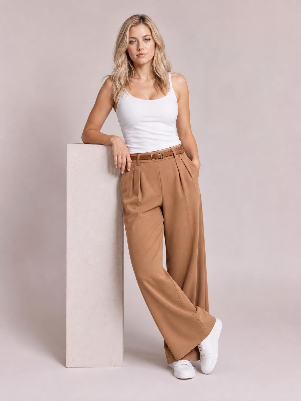Model poses in camel pleated wide-leg pants with a high waist, front pockets, and relaxed flowing legs, paired with a white tank top.