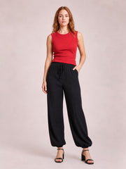 A model wears black drawstring jogger pants with a relaxed wide-leg silhouette, tied waist cord, side pockets, and gathered elastic cuffs, standing with one hand in a pocket.