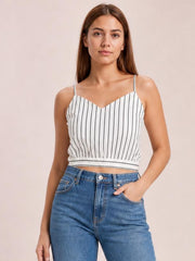 A woman models a white-and-black striped cropped cami with a V-neck, slim spaghetti straps, sleeveless construction, and a fitted silhouette, posing with one hand in her jeans pocket.