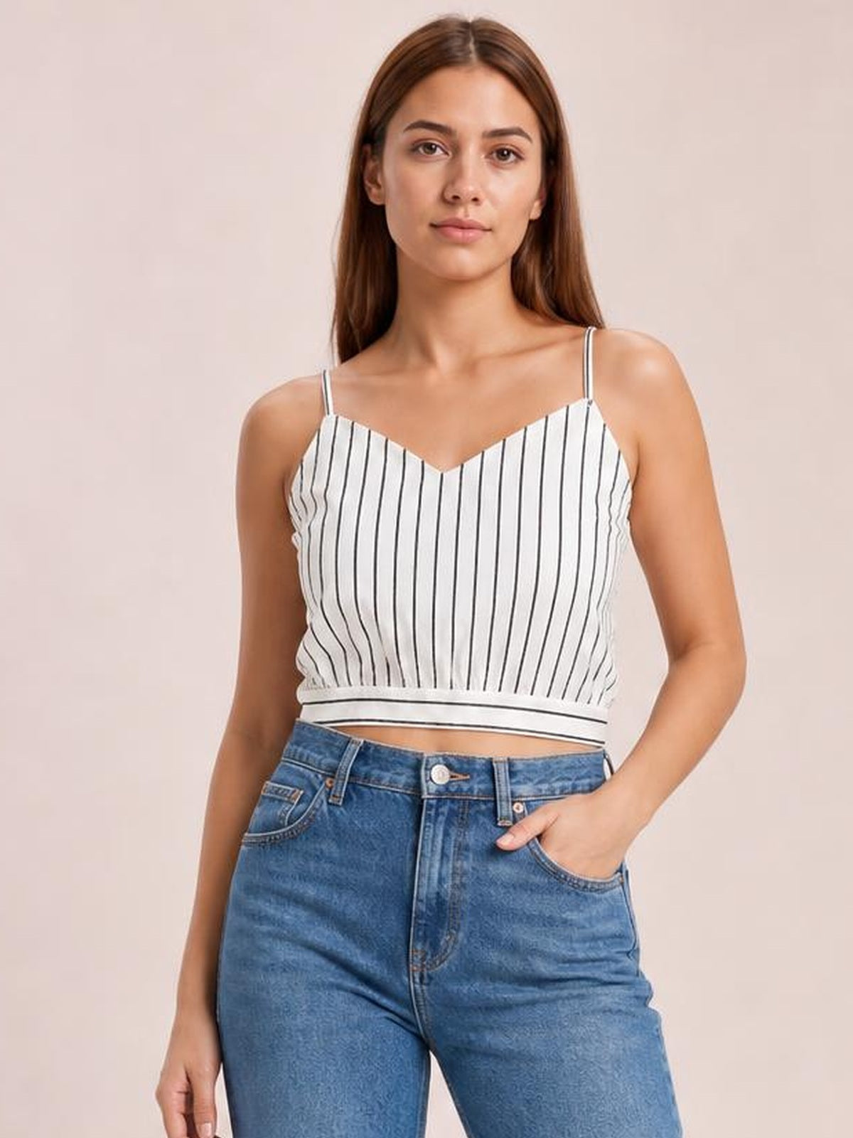 A woman models a white-and-black striped cropped cami with a V-neck, slim spaghetti straps, sleeveless construction, and a fitted silhouette, posing with one hand in her jeans pocket.