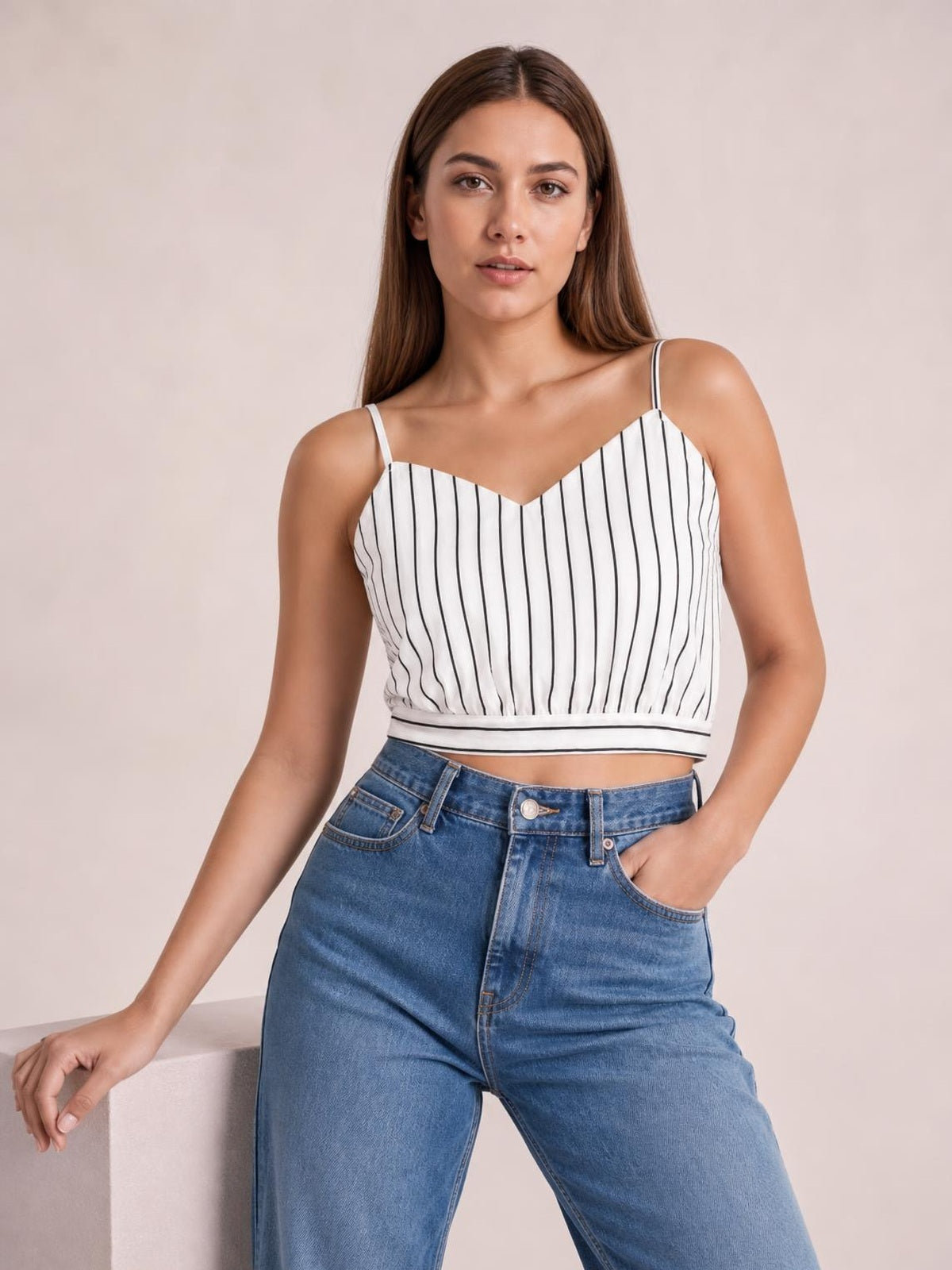 Model wears a white striped tie-back cropped cami with a V-neck, sleeveless spaghetti straps, and a fitted banded hem while posing with one hand in her jeans pocket.