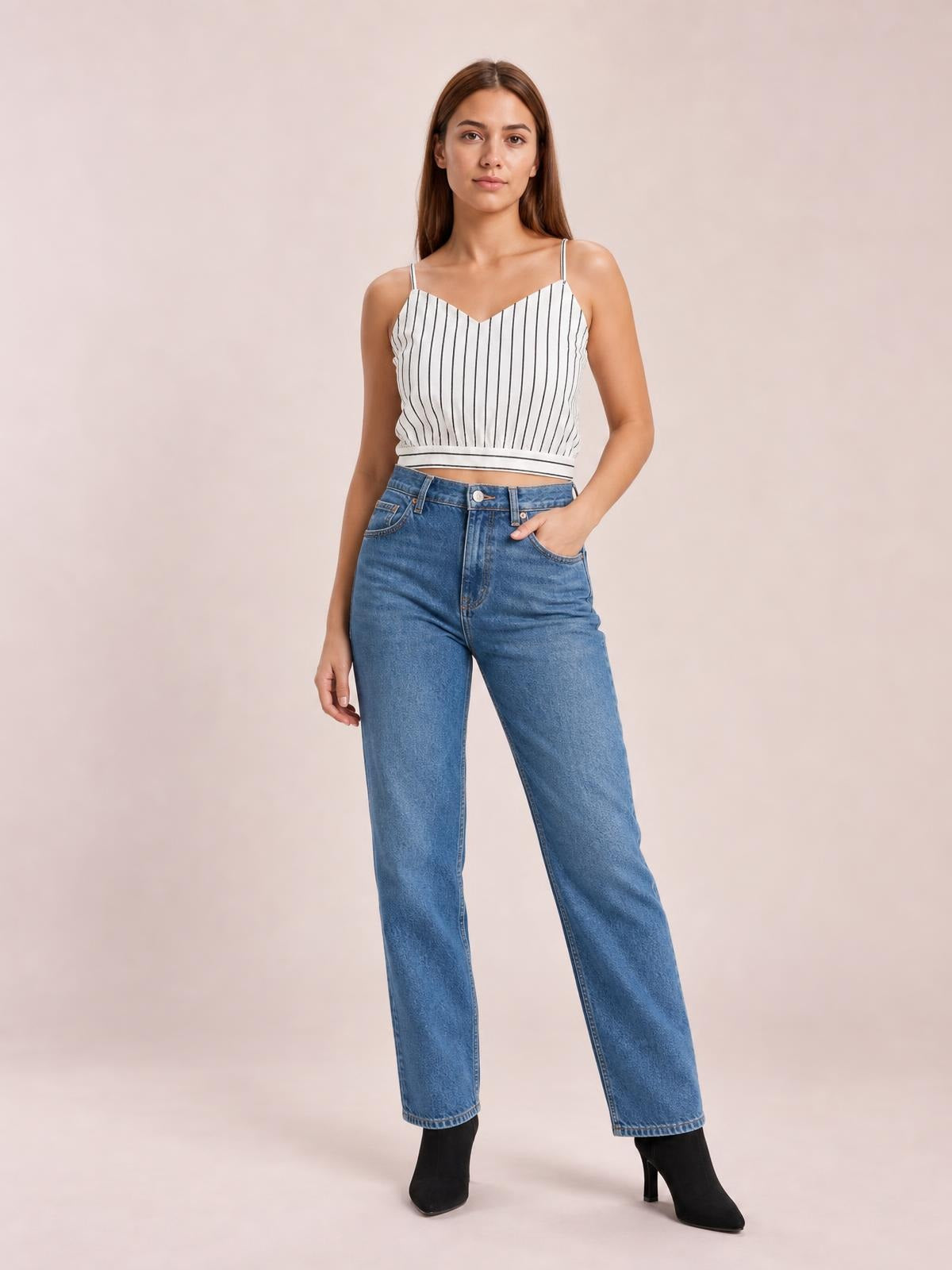 Model poses with one hand in her pocket wearing a white striped cropped cami with a V-neck, spaghetti straps, and a defined waist.