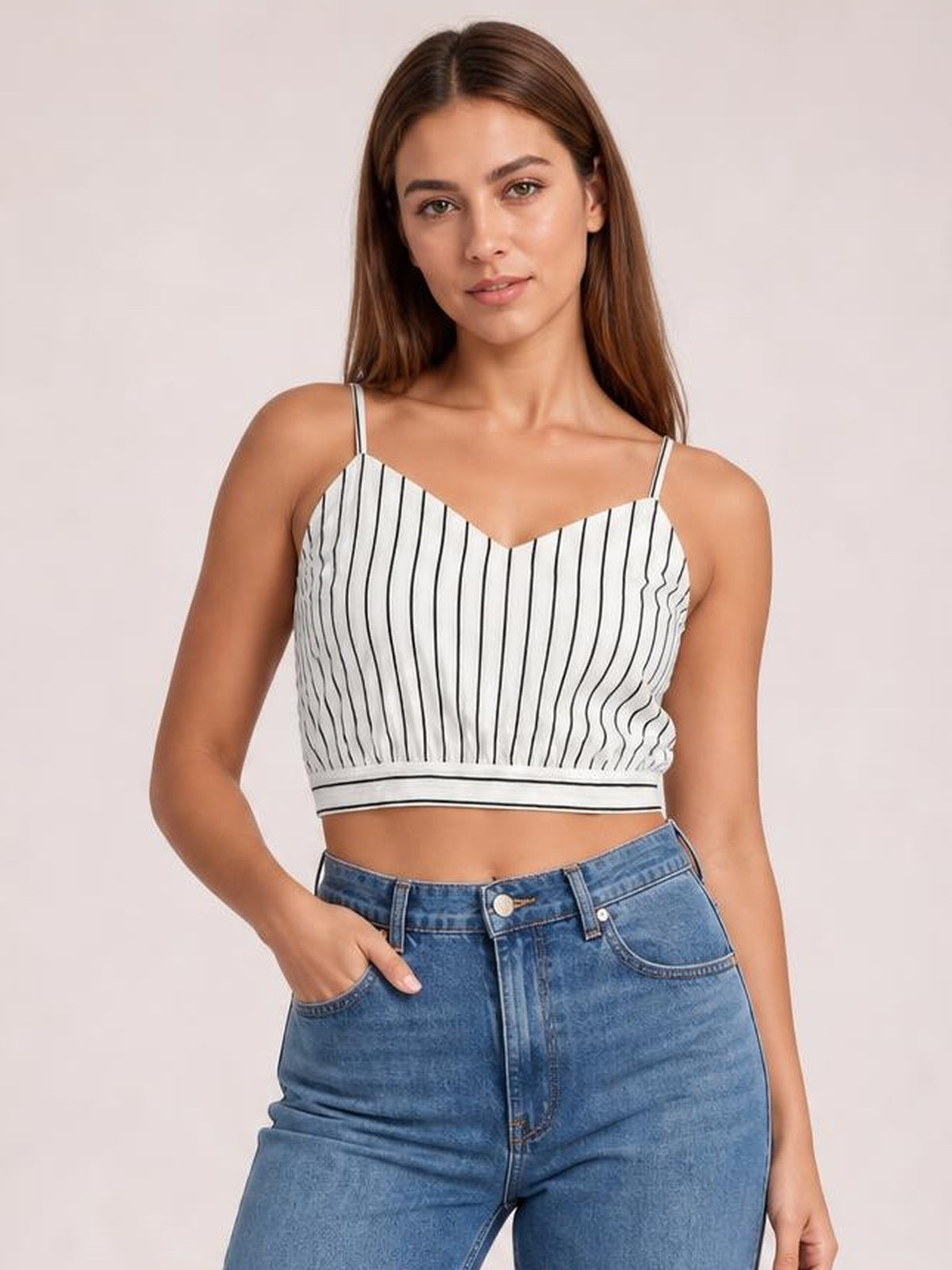 Model wearing a white striped cropped cami with black vertical lines, V-neckline, spaghetti straps, fitted silhouette, and a hand resting in her jeans pocket.