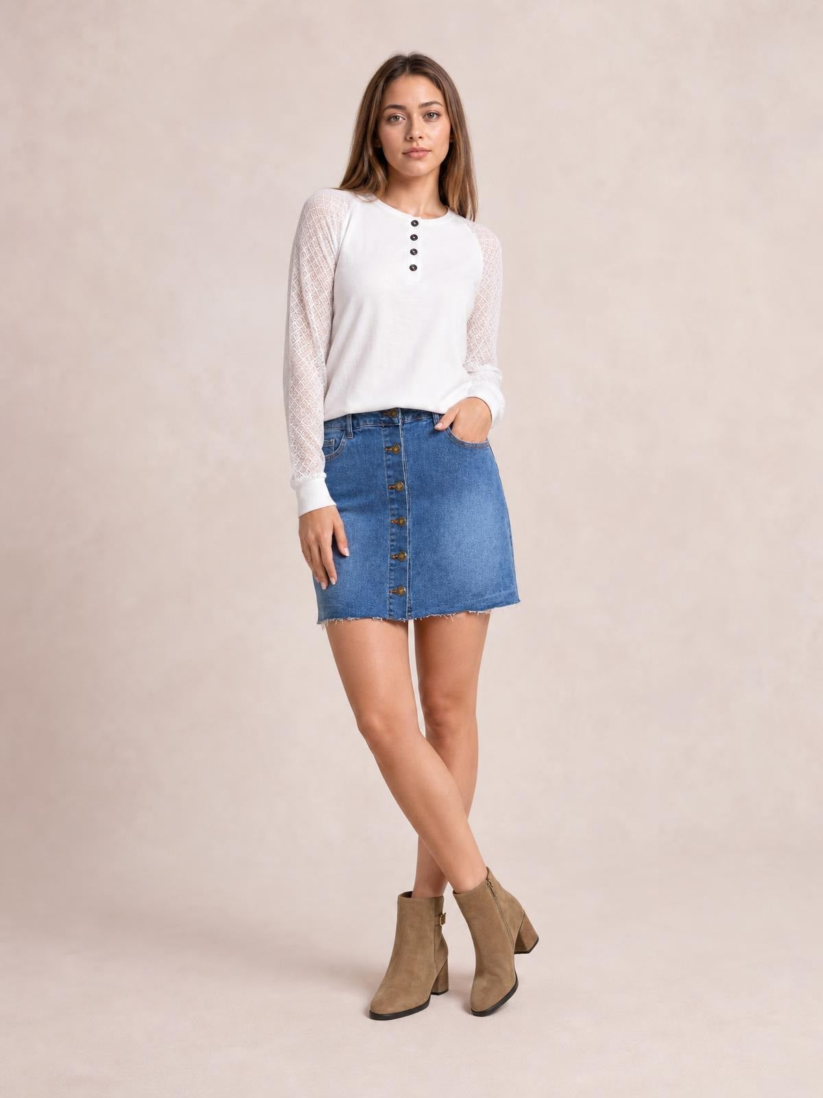 A model poses with crossed legs and one hand in a pocket, wearing a blue denim mini skirt with a front button closure, side pockets, and a raw hem.