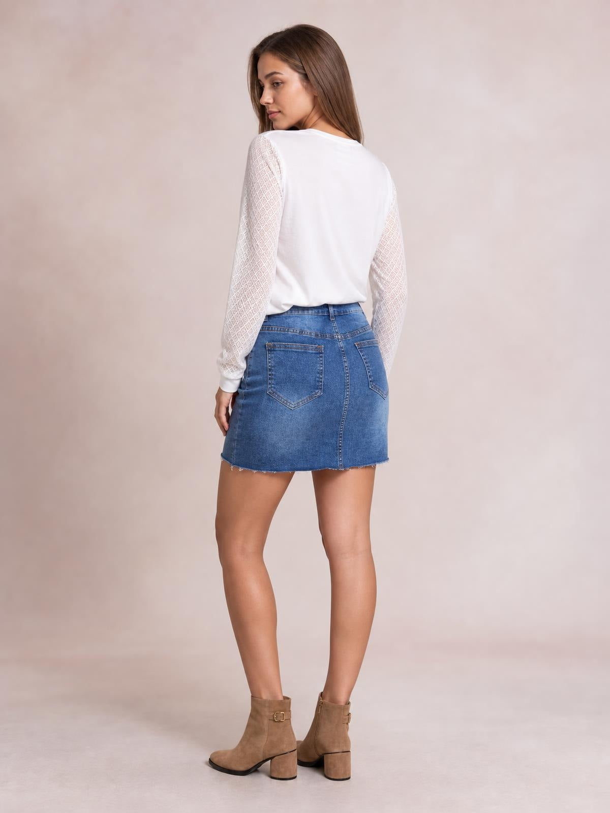 Model looks over her shoulder while wearing a blue denim skirt with a fitted silhouette, rear patch pockets, structured seams, and a raw mini hem.