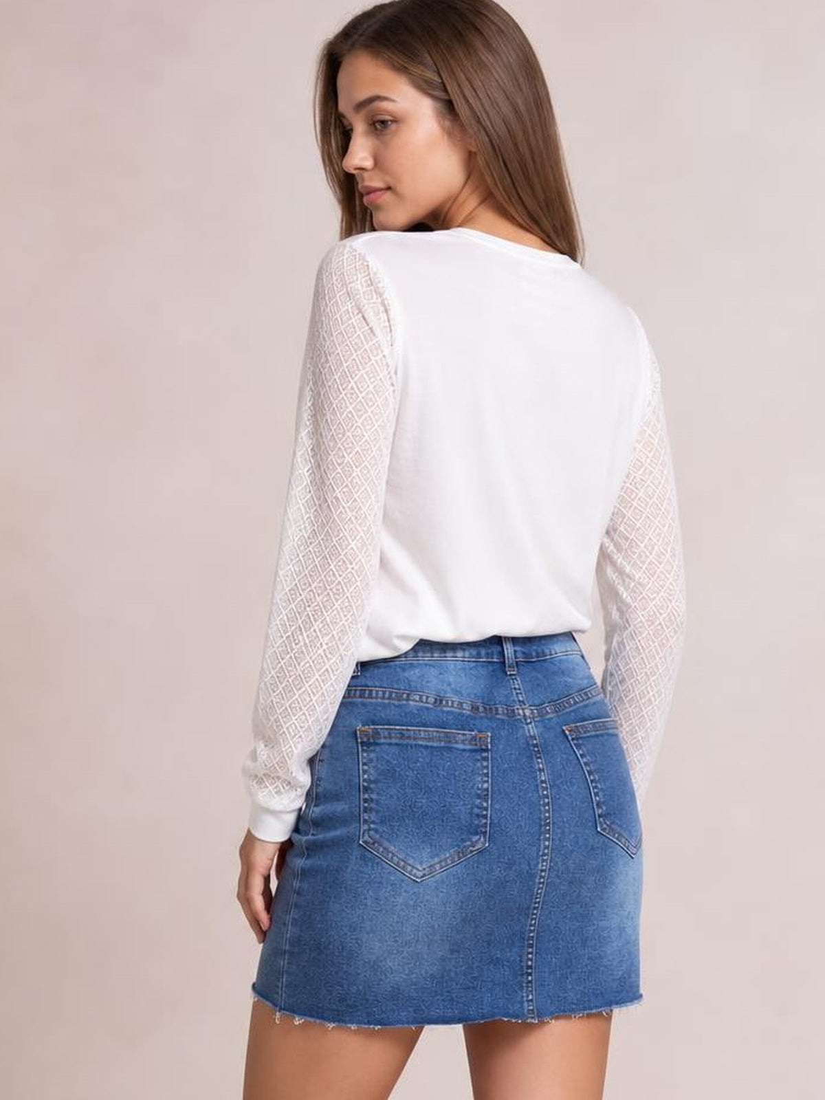 A model looks over her shoulder in a blue pocketed denim skirt with a fitted silhouette, belt loops, back patch pockets, and a raw mini hem.