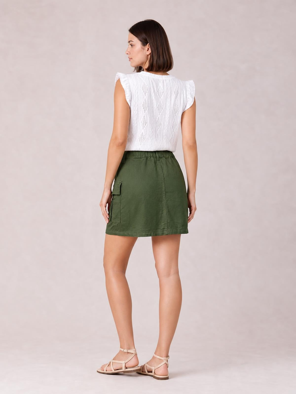 A model poses in a back view wearing an olive green pocketed denim mini skirt with a straight silhouette, elastic waistband, and center seam.