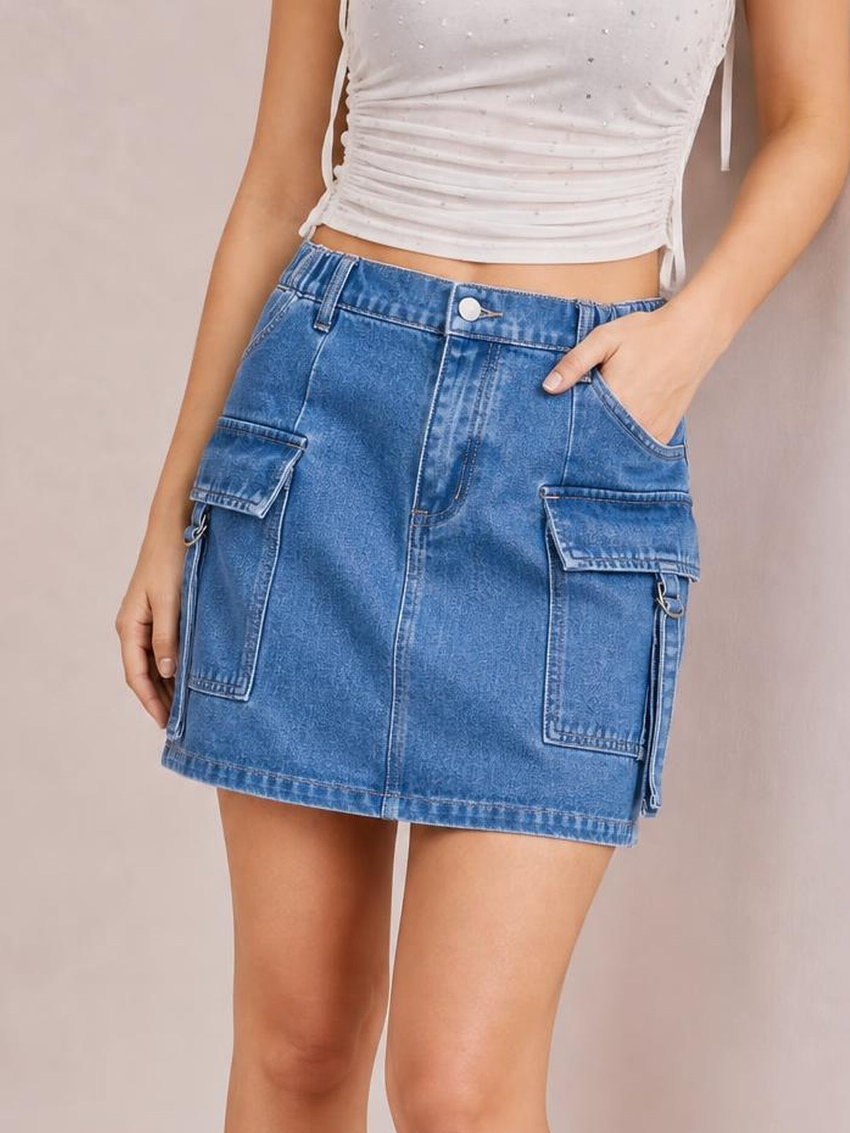 Model wearing a blue buttoned denim skirt with a straight mini silhouette, front seam detailing, and multiple flap pockets, posing with one hand in a pocket.