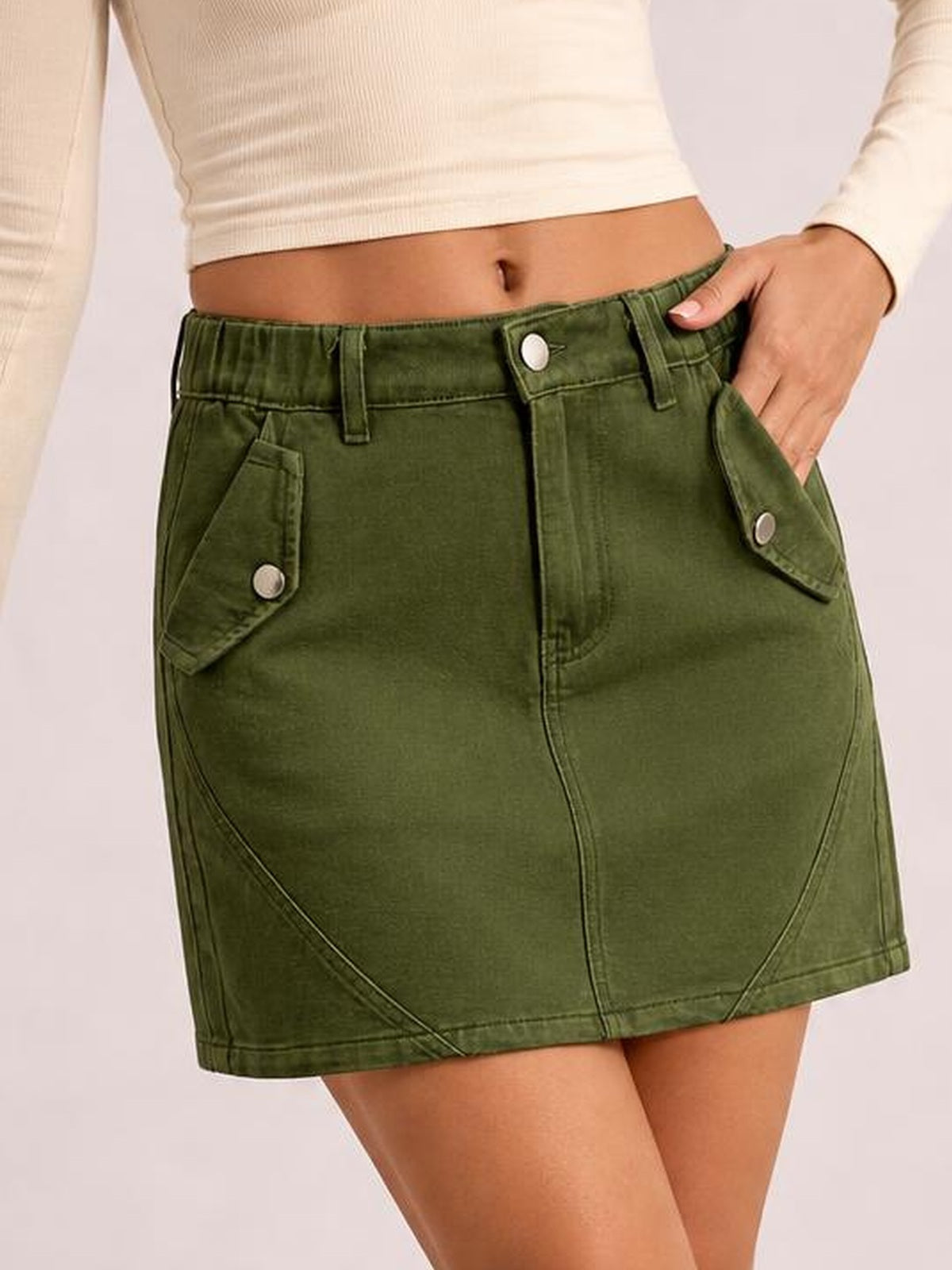 Model wearing a green pocketed buttoned mini denim skirt with a fitted silhouette, front seam construction, and flap pockets while resting a hand in one pocket.
