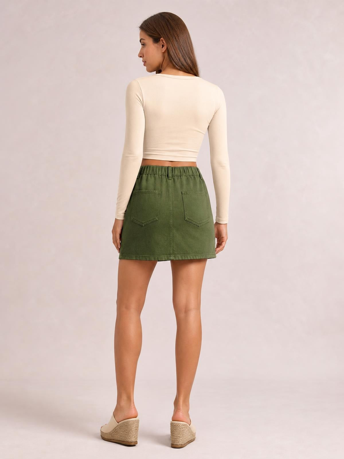 Model shown from behind wearing a green mini denim skirt with two patch back pockets, an elasticized waistband, straight silhouette, and stitched hem.
