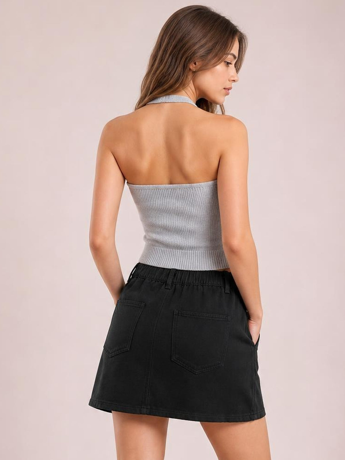 A model poses from the back in a black mini denim skirt with two patch pockets, belt loops, and a gently flared silhouette, hands in pockets.