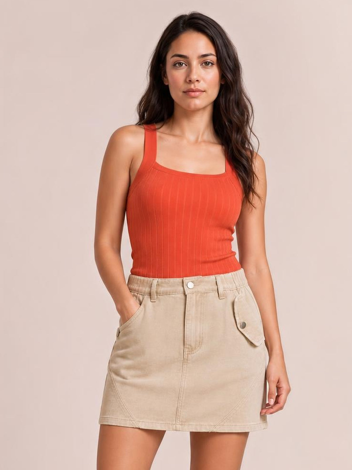 A model stands with one hand in a pocket, wearing a beige mini denim skirt with a straight silhouette, front button closure, flap pocket, and visible topstitching.