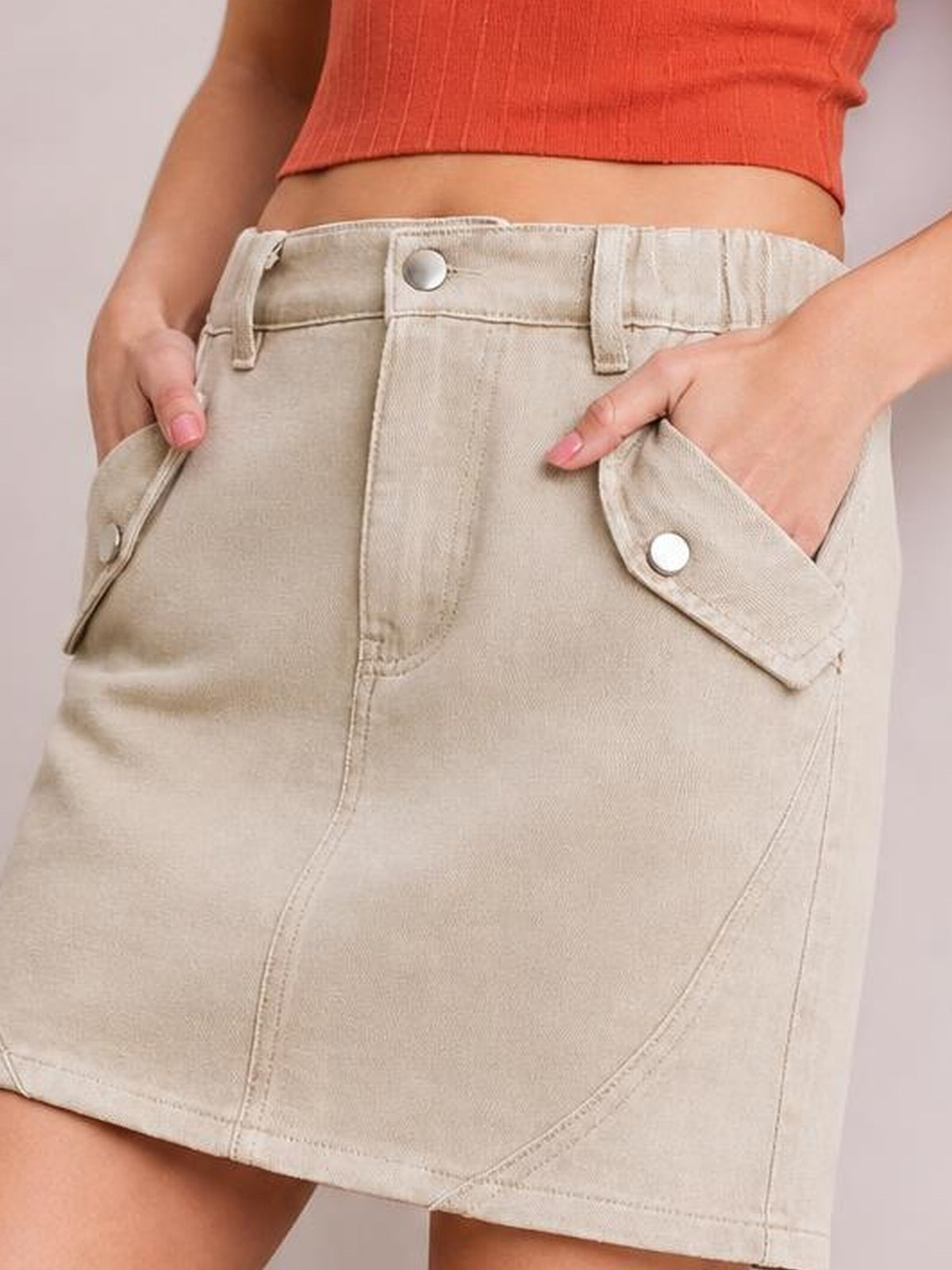 Model wearing a light beige pocketed buttoned mini denim skirt with belt loops, front seams, flap pockets, and a straight silhouette.