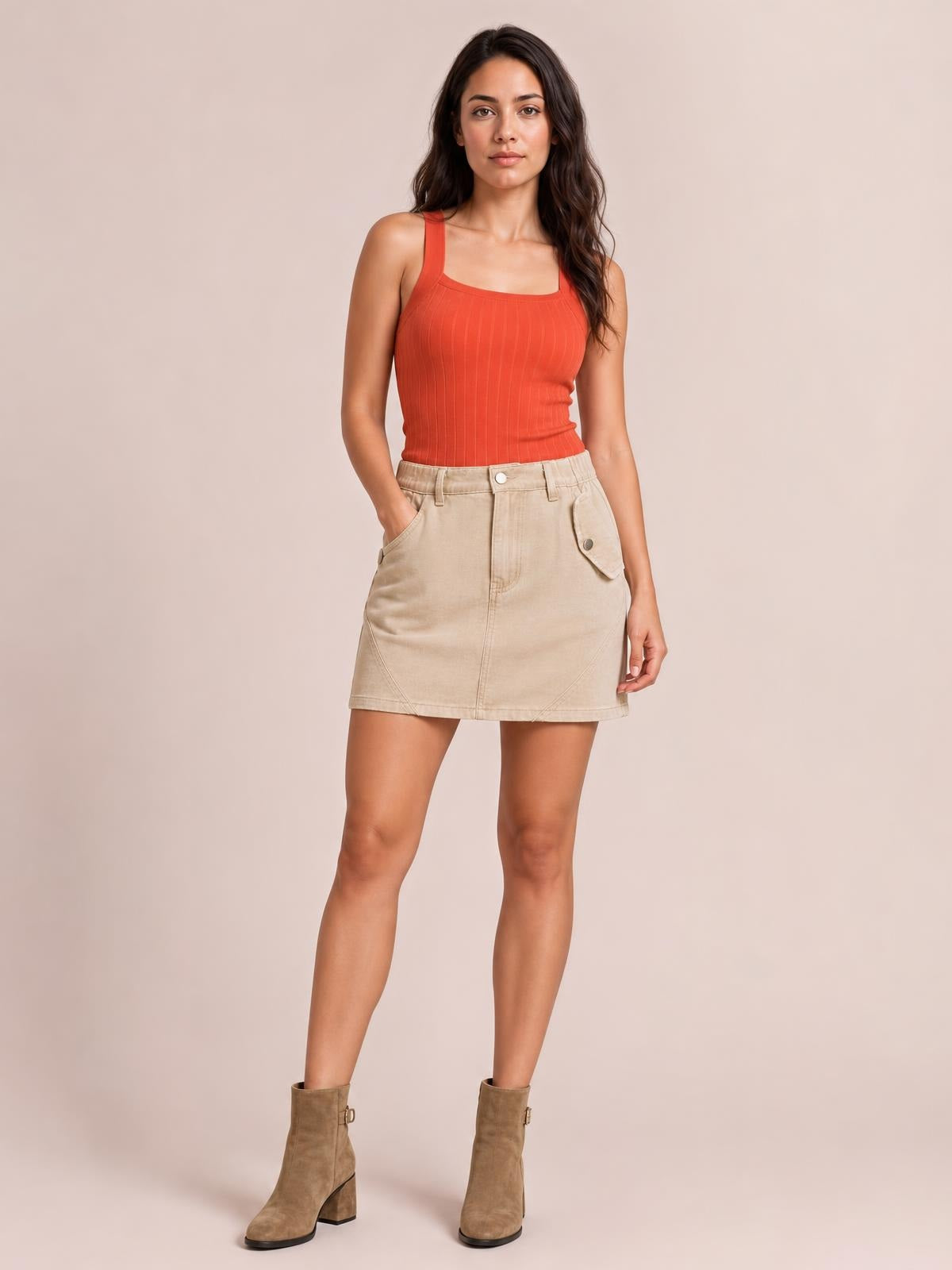 A model wears a beige pocketed buttoned mini skirt with belt loops, front seams, and a straight silhouette, standing with one hand in a pocket.