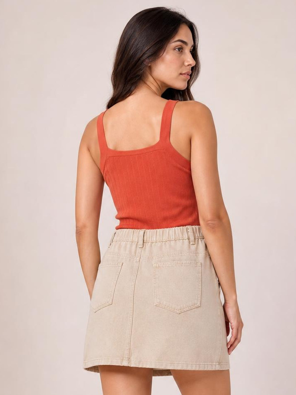 Model looks over her shoulder wearing a beige mini denim skirt with a straight silhouette, belt loops, stitched back pockets, and a clean hem.