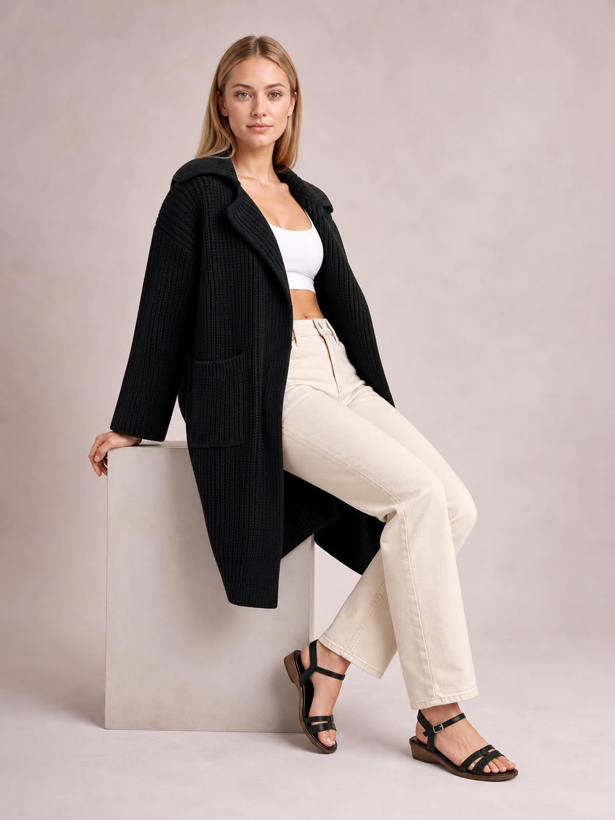 Model seated in a relaxed pose wears a black pocketed collared neck cardigan with dropped shoulders, textured knit, long sleeves, and an elongated silhouette.