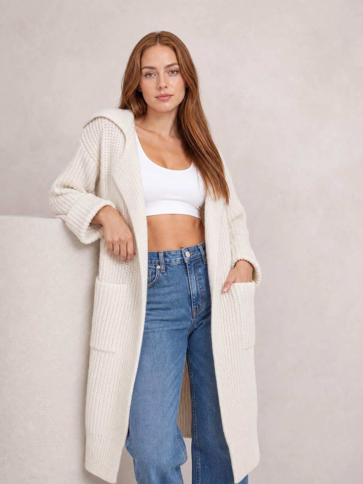 A model poses in a relaxed ivory longline cardigan with dropped shoulders, chunky knit texture, wide cuffs, and large patch pockets, worn open over a white top.