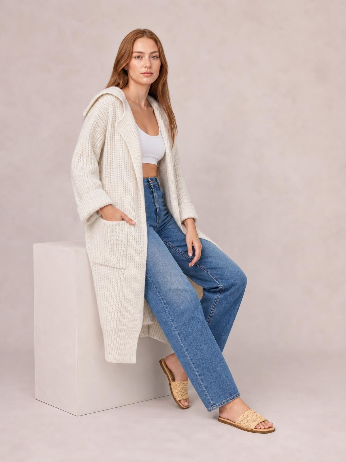 Model sits with one hand in the pocket of a cream dropped-shoulder cardigan featuring a collared neckline, open front, long sleeves, and relaxed long silhouette.