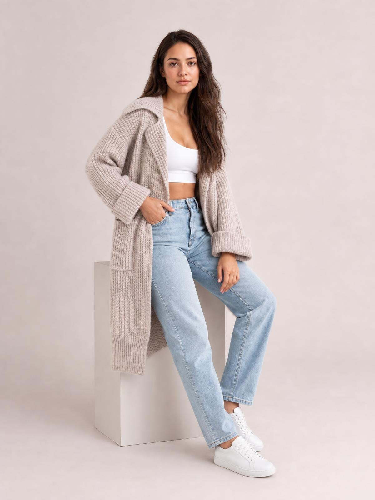 Model seated in a relaxed pose wears a light taupe pocketed collared neck cardigan with dropped shoulders, chunky ribbed knit, long silhouette, and wide cuffs.