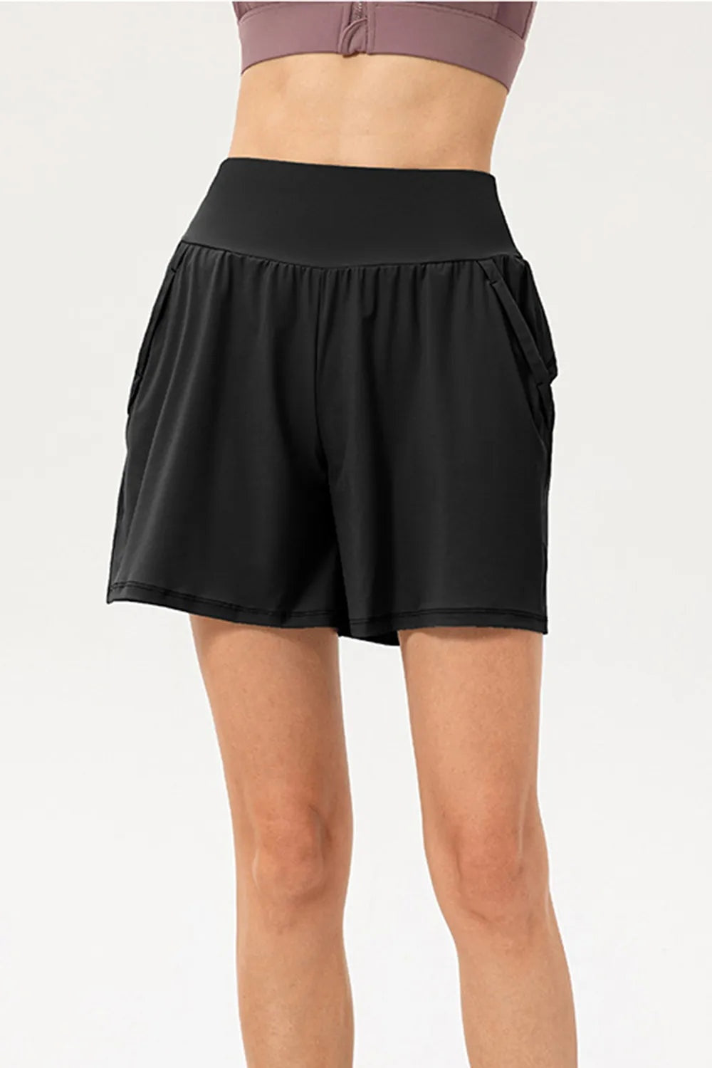 A standing model wears black active shorts with a wide elastic waistband, relaxed flowy silhouette, side pockets, and neatly stitched hems.