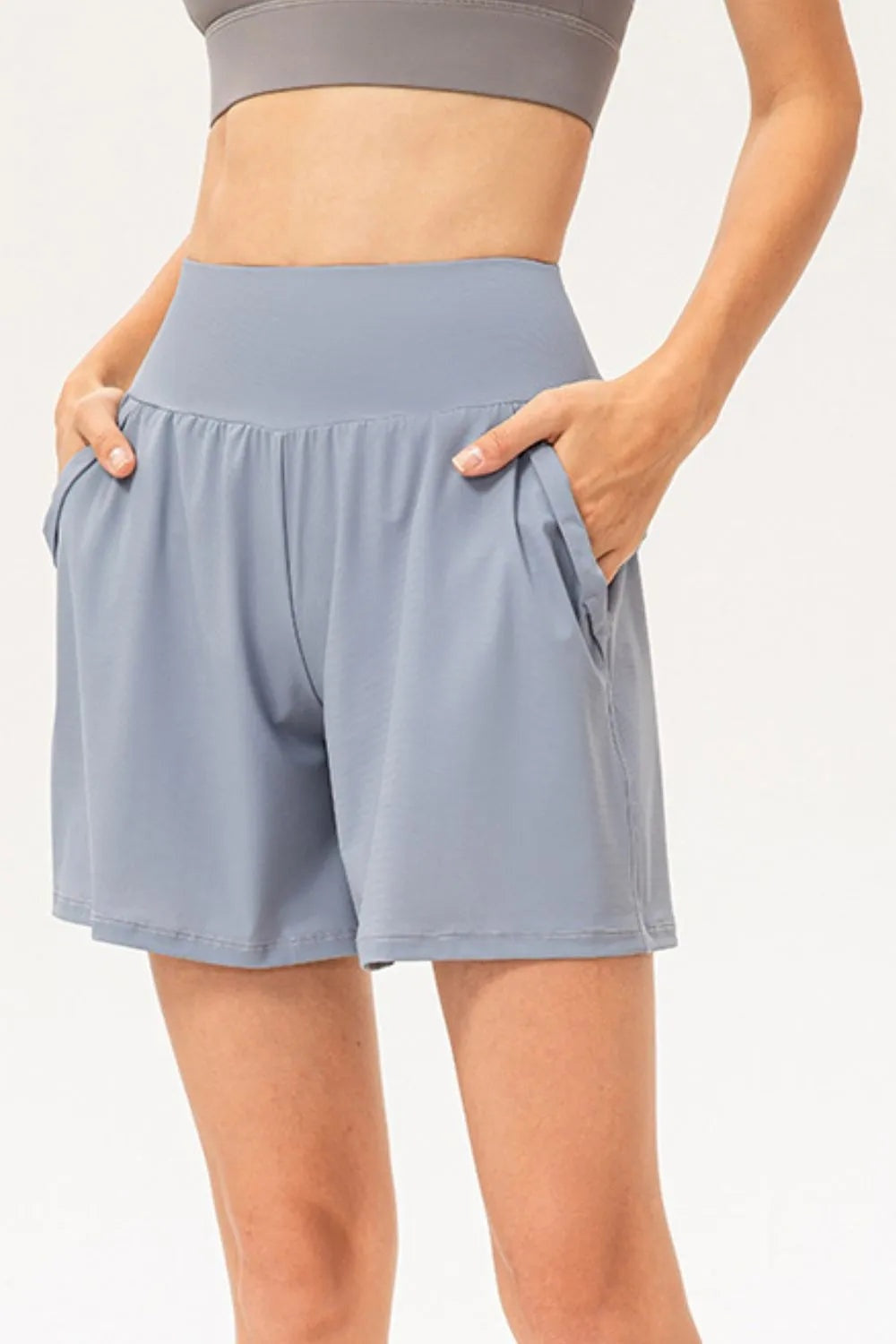 Model wears blue-gray pocketed active shorts with a wide elastic waistband, relaxed silhouette, and hands tucked into the side pockets.