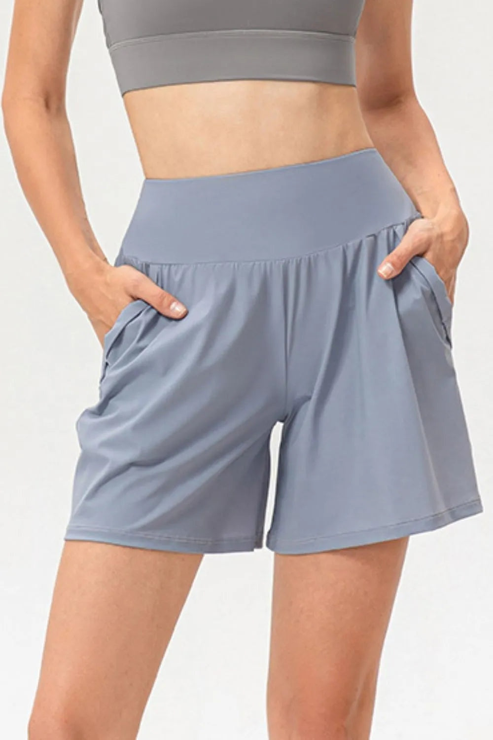 A model stands with hands in the side pockets, wearing slate blue active shorts with an elastic waist, relaxed wide-leg silhouette, and stitched hems.