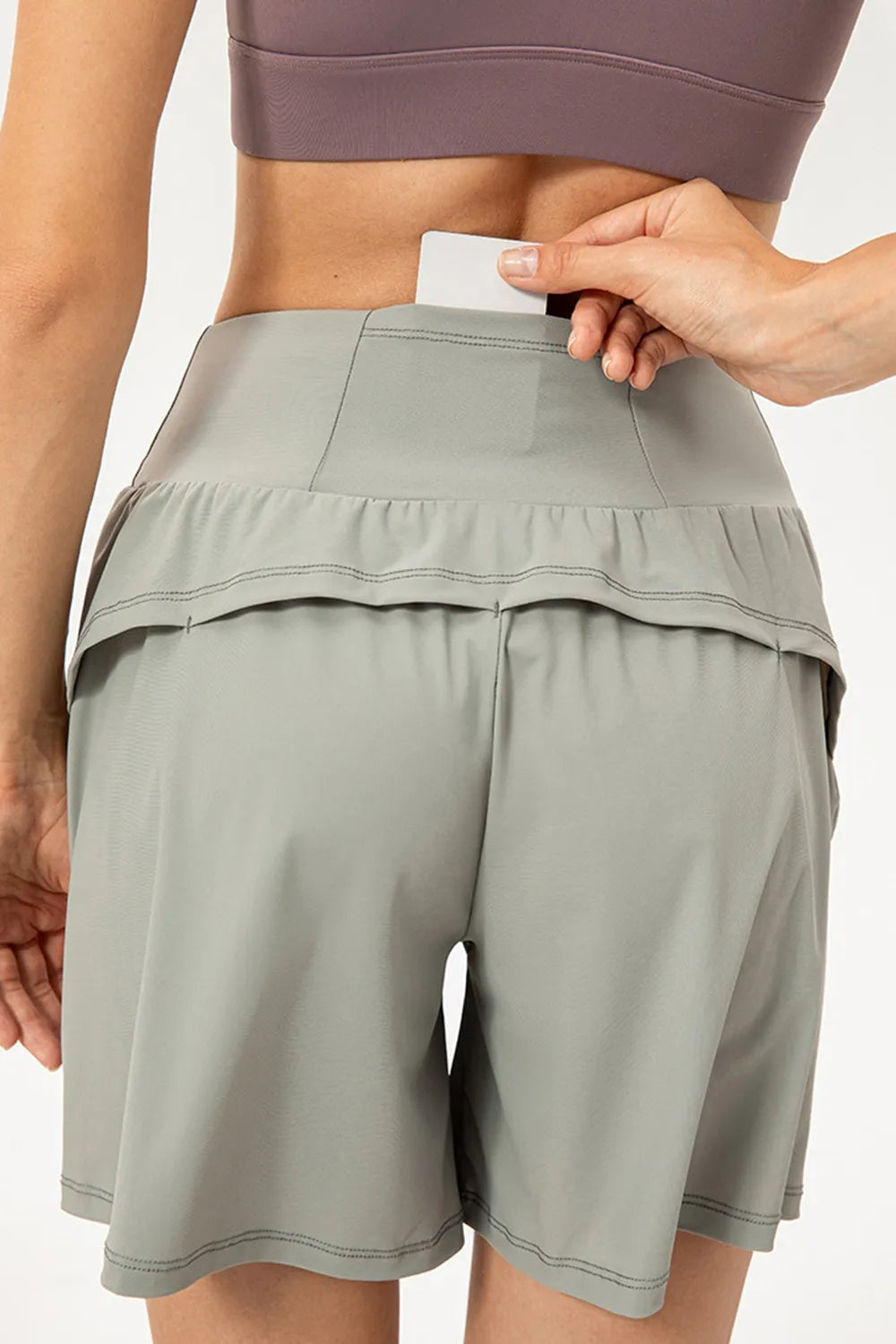 A model wears light gray pocketed active shorts with an elastic waist, relaxed mid-thigh silhouette, gathered overlay, and double-stitched hems.