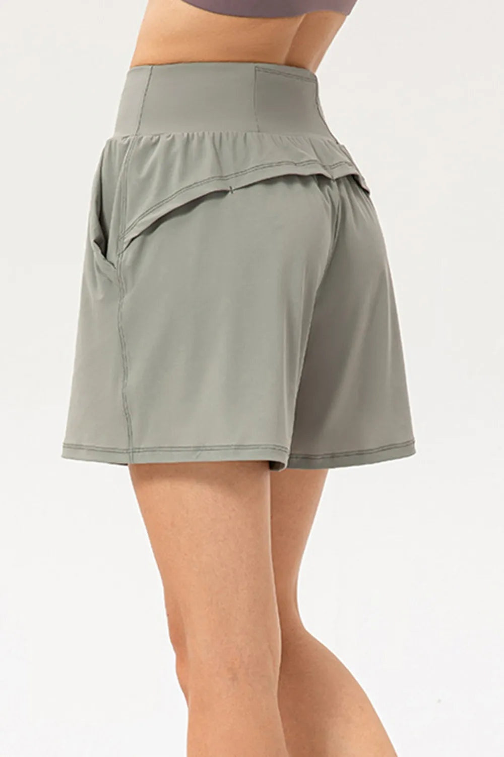Model wears sage green pocketed active shorts with a wide elastic waist, relaxed flared silhouette, curved overlay panel, and visible contrast stitching.