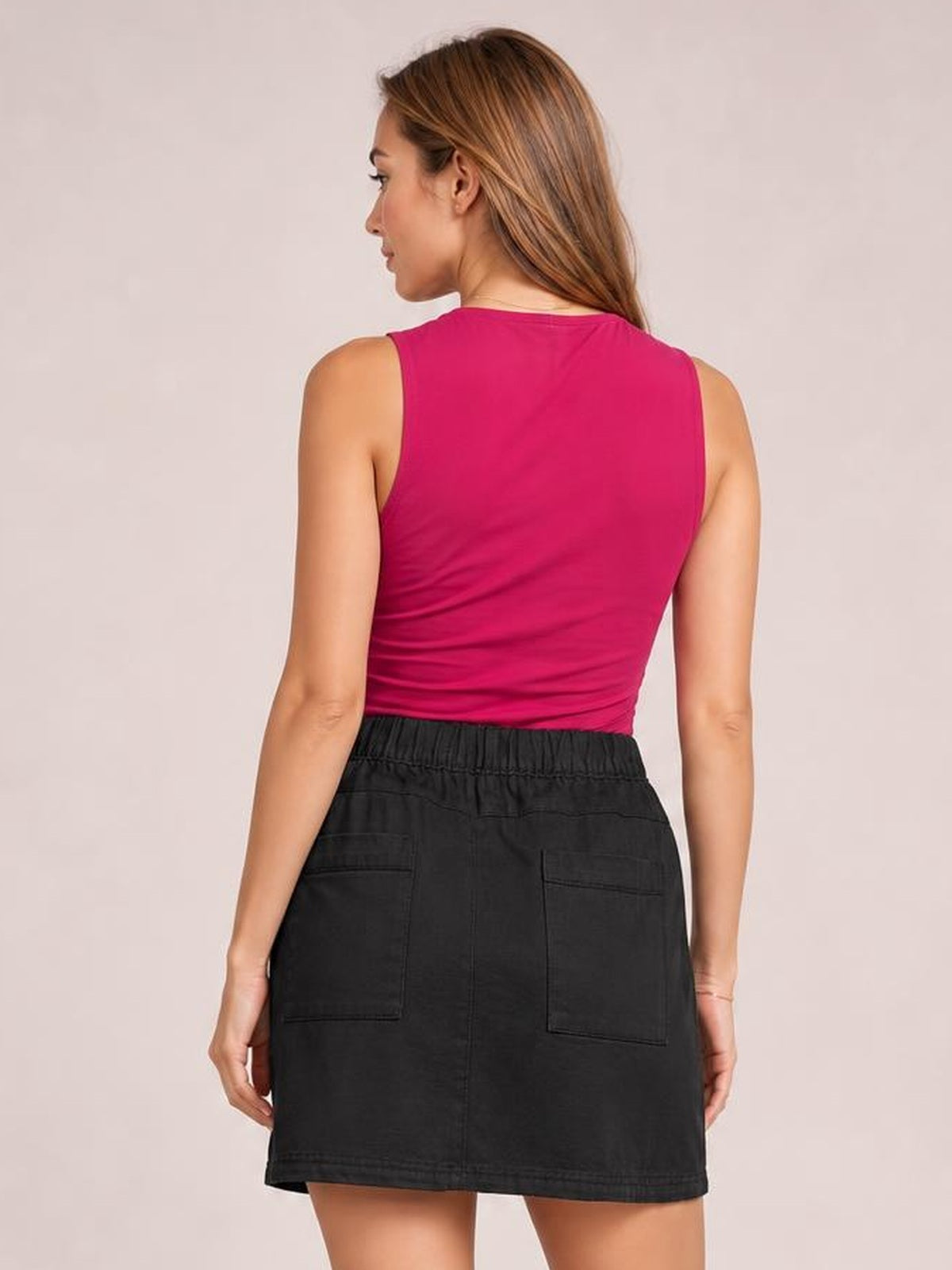 A model shown from behind wears a black denim skirt with an elastic waistband, stitched back patch pockets, and a straight above-knee silhouette.