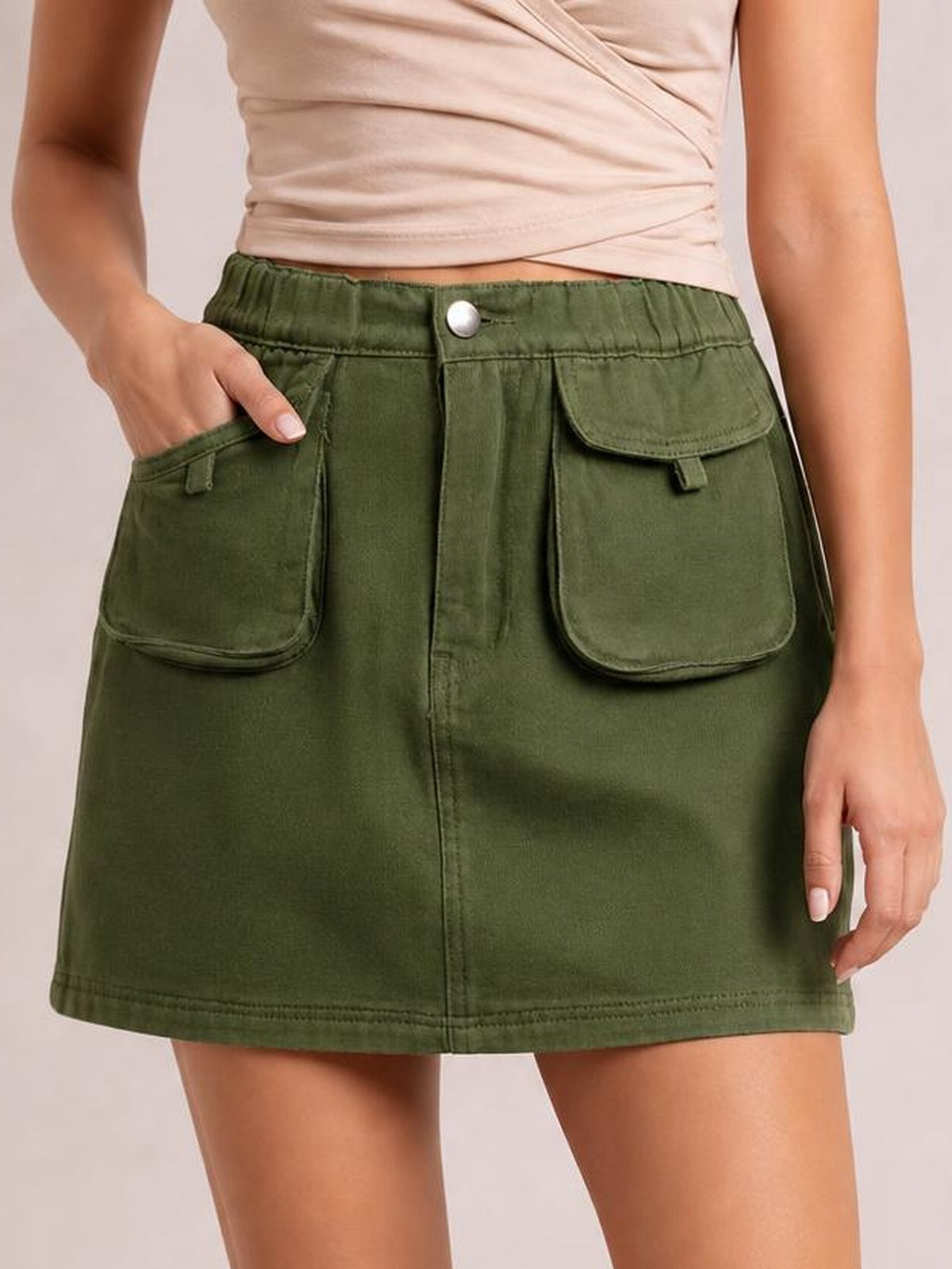A model wears an olive-green denim skirt with an elastic waist, button closure, front patch pockets, and a straight mini silhouette, with one hand tucked inside.