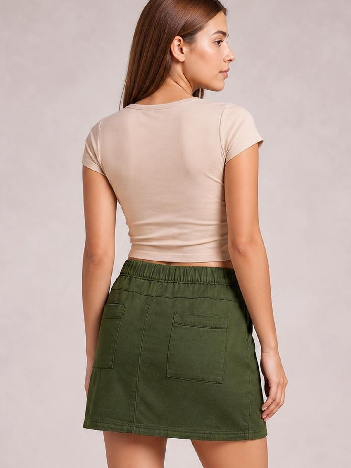 A model faces away wearing an olive green denim skirt with an elastic waistband, back patch pockets, stitched panel seams, and a straight mini silhouette.