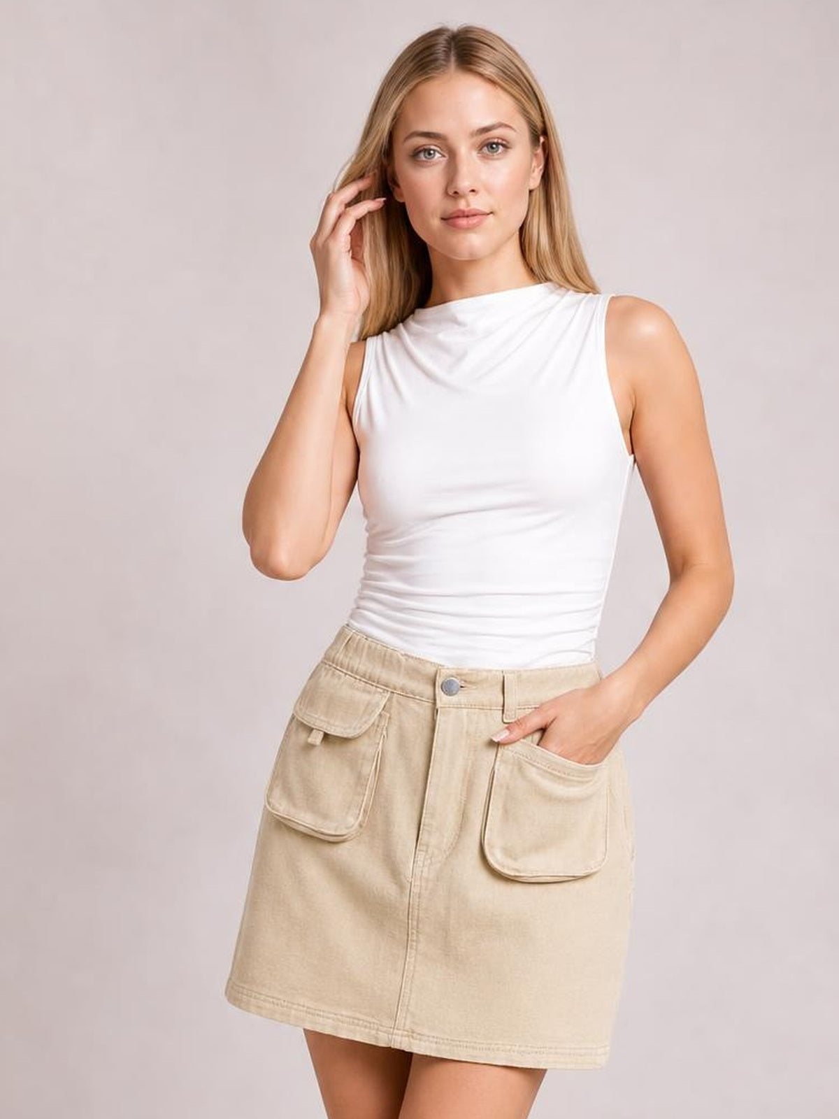 A model poses in a beige denim skirt with an elastic waist, button closure, large patch pockets, and a straight mini silhouette, resting one hand in a pocket.
