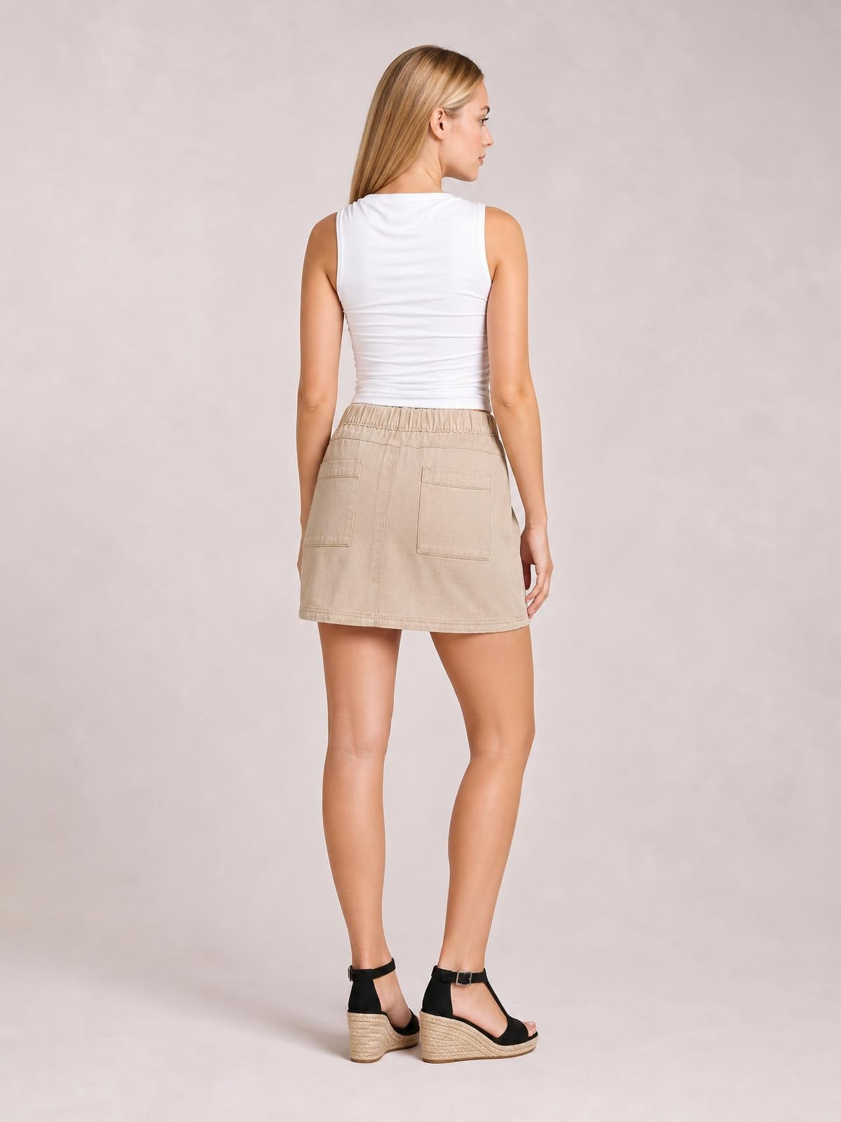 Model shown from behind wears a beige denim skirt with an elastic waist, two back patch pockets, visible stitching, and a straight mini silhouette.