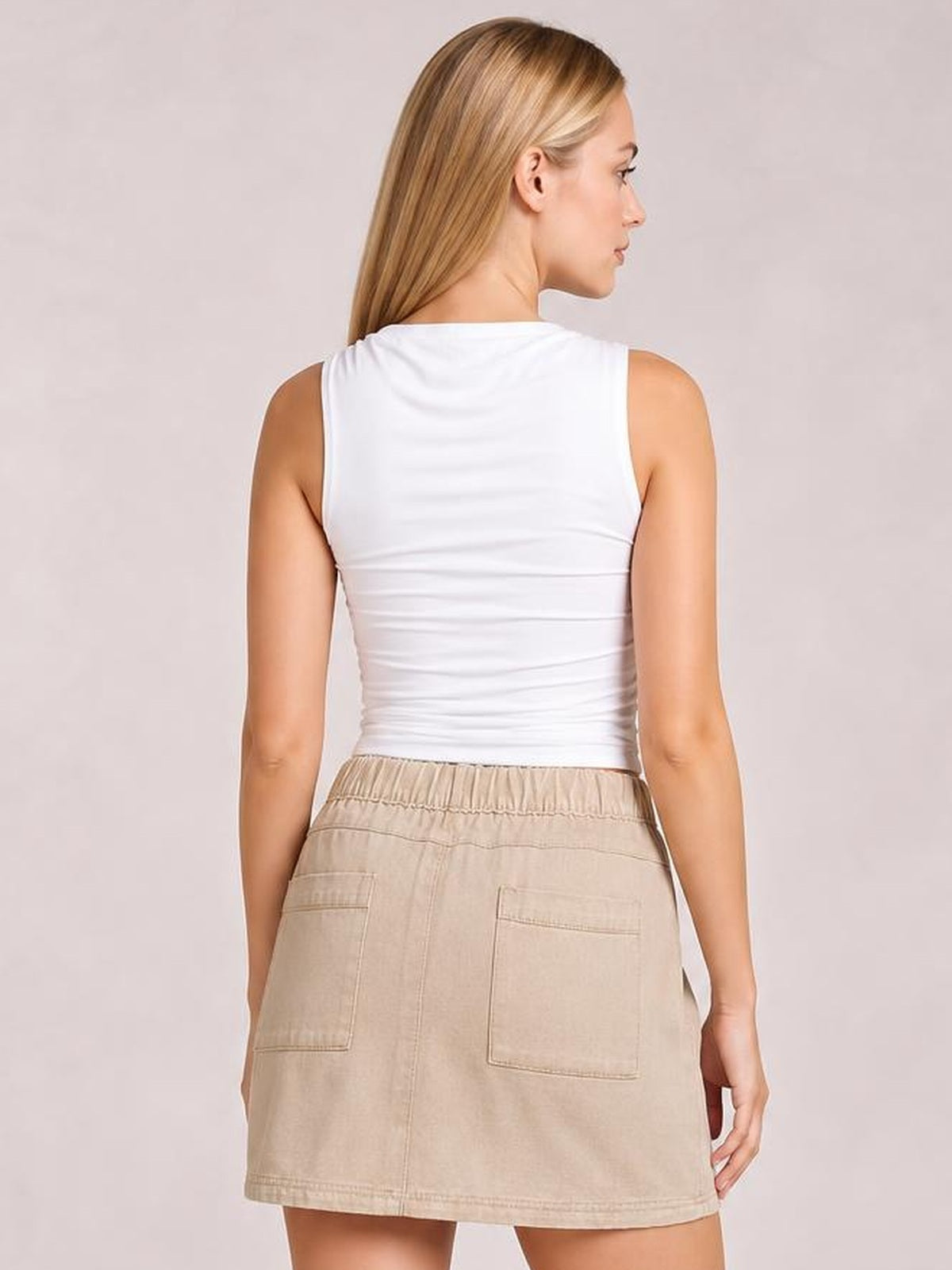 A model poses in profile wearing a beige denim mini skirt with an elastic waist, straight silhouette, center seam, and two large patch pockets on the back.