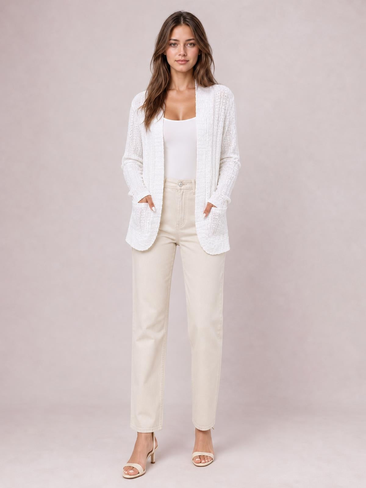 Model wearing a white eyelet open-front cover-up with long sleeves, rounded hem, and front pockets over a fitted top, posing with hands in pockets.