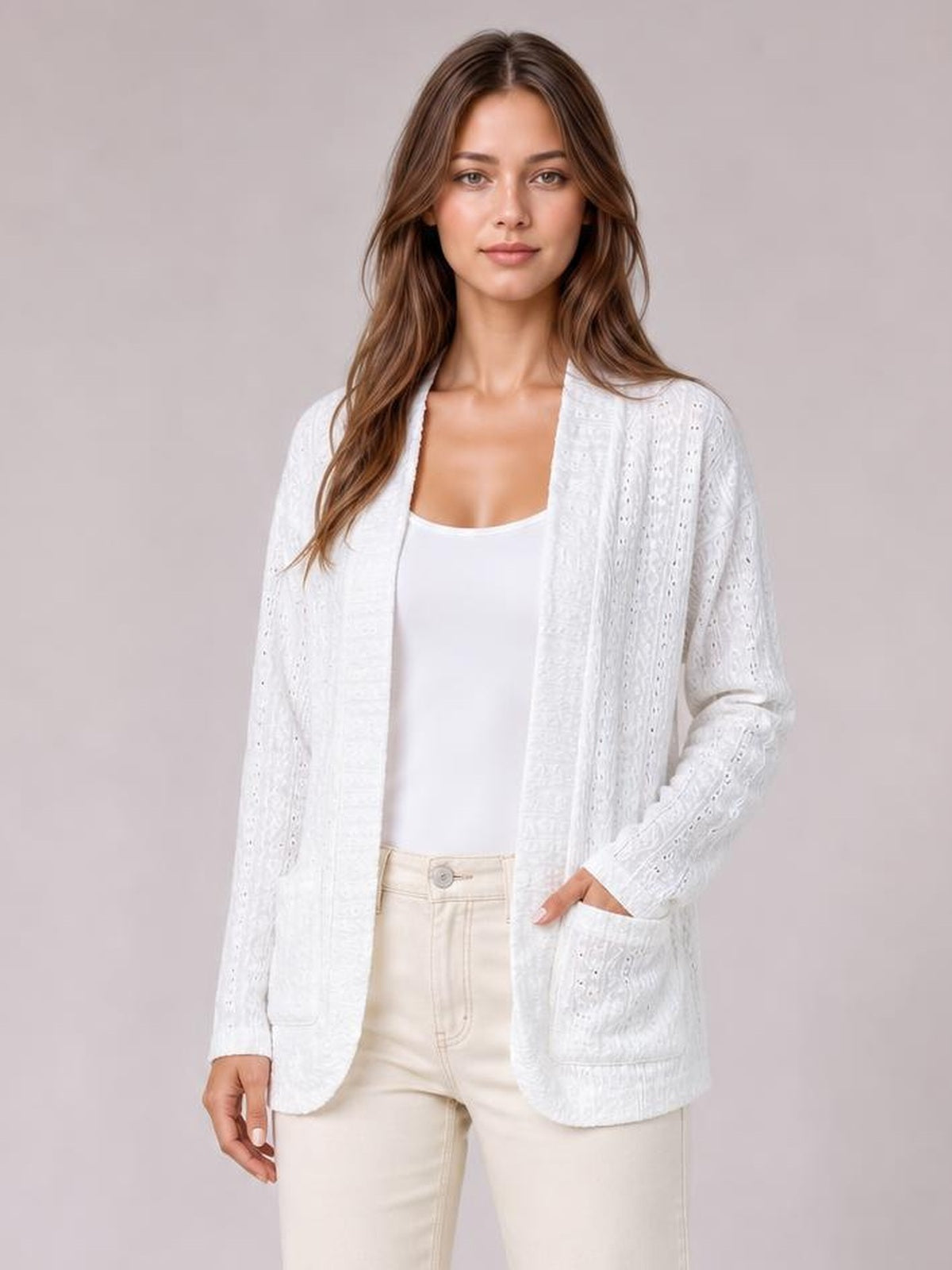 Model stands with one hand in a pocket, wearing a white eyelet open-front cover-up with long sleeves, a relaxed silhouette, and rounded hem over a white top.