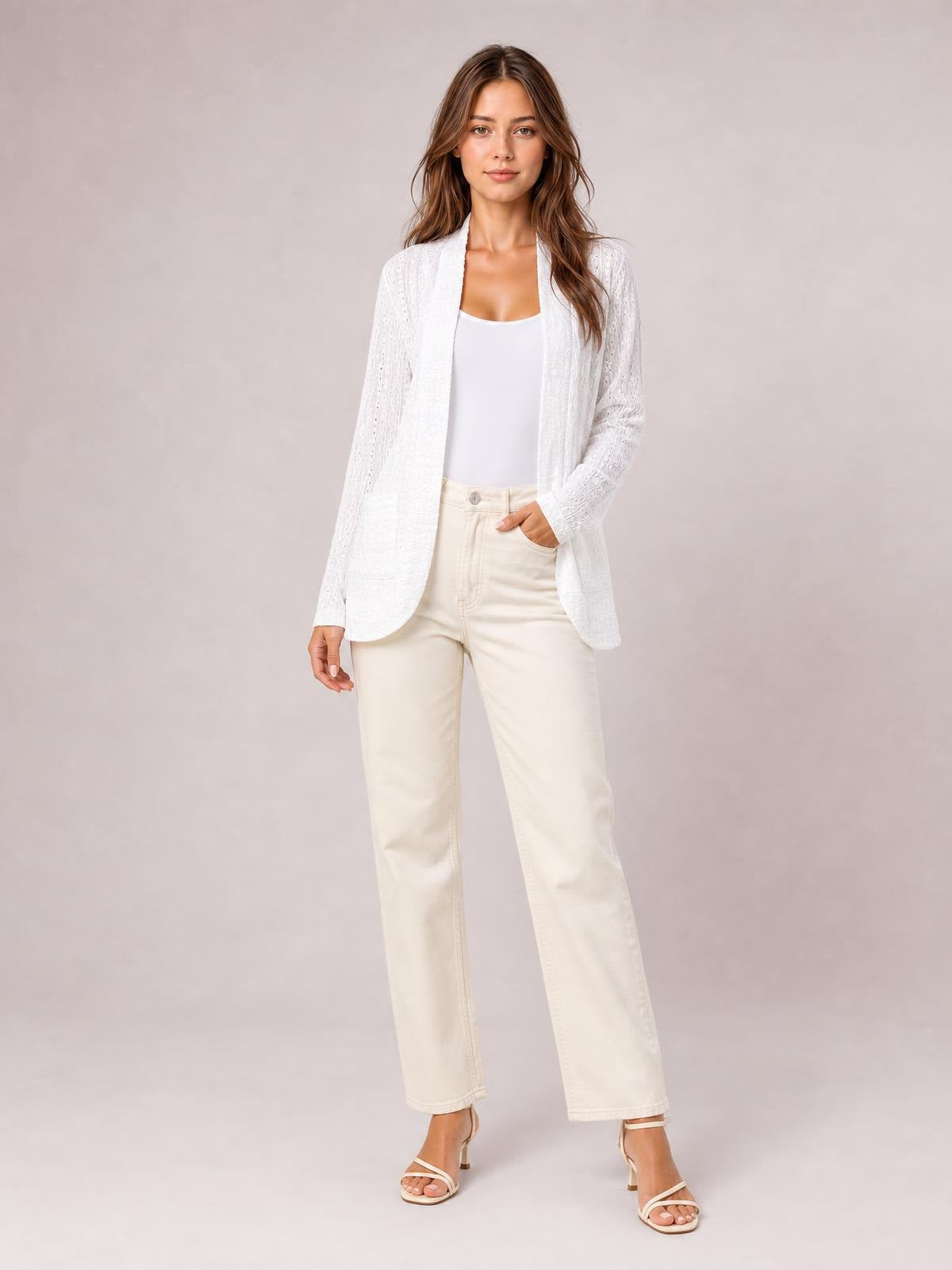 A model stands in a white eyelet open-front cover-up with long sleeves, curved hem, relaxed silhouette, and visible lower patch pockets over a tank top.