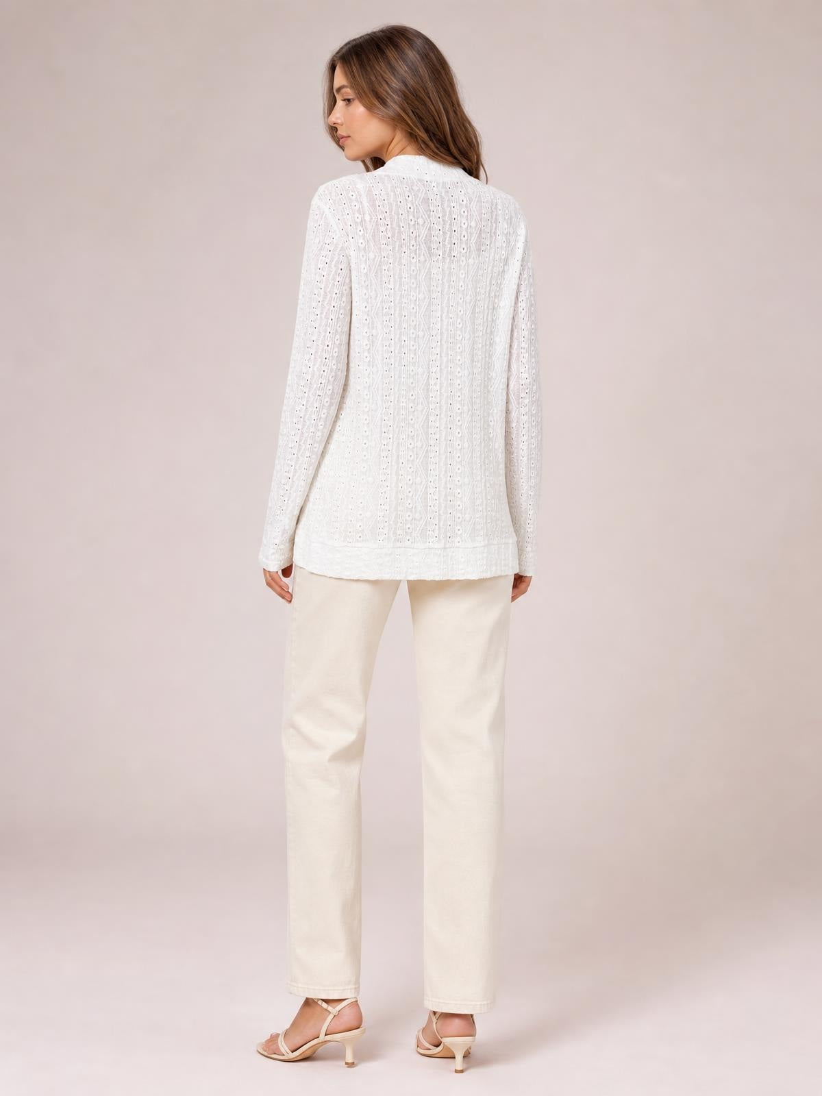 A model stands turned slightly away, showcasing a white eyelet cover-up with long sleeves, a relaxed straight silhouette, and a softly finished hem.