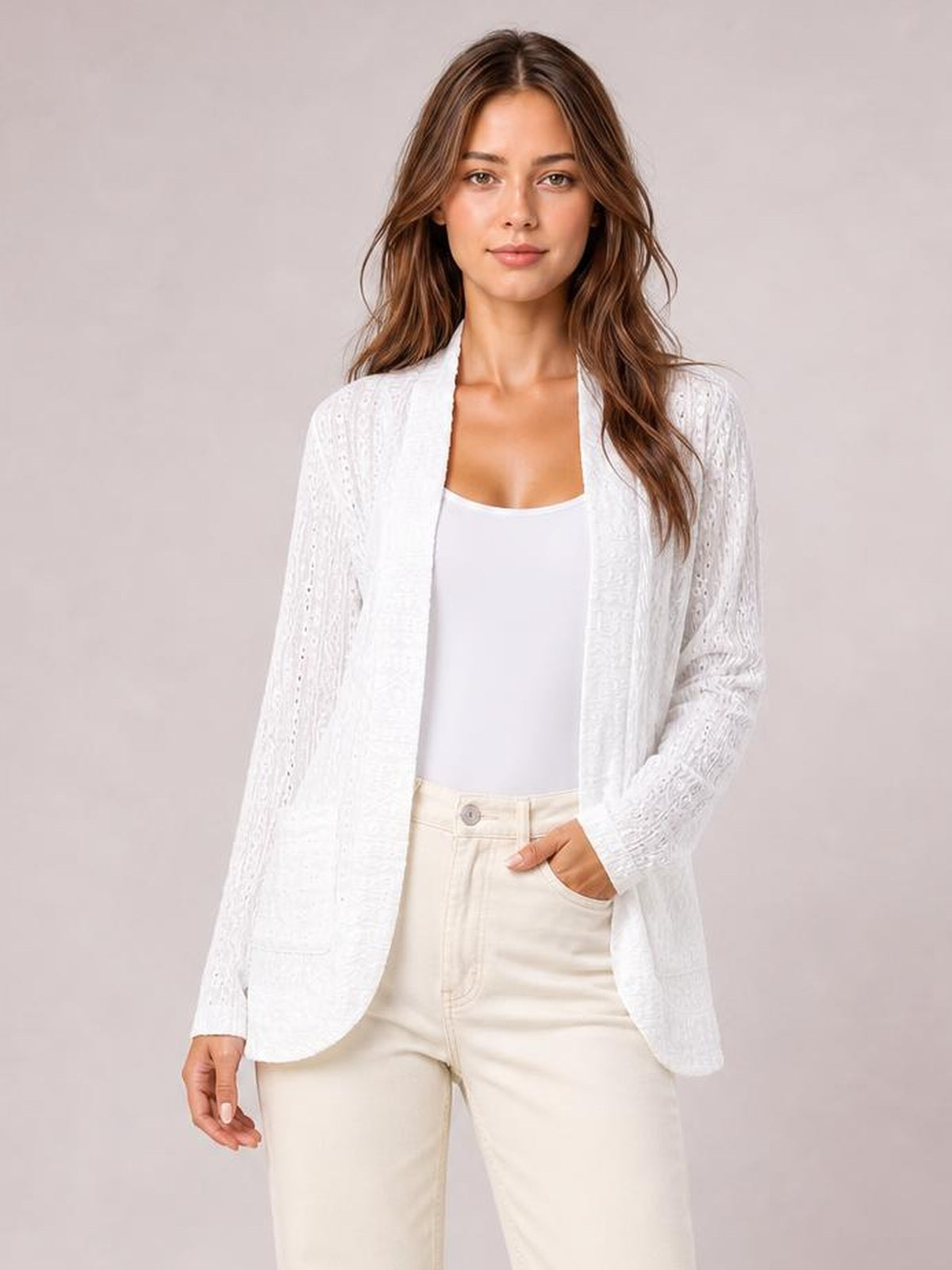 Model wears a white eyelet open front cover up with long sleeves, curved hem, relaxed silhouette, and visible front pockets over a white top.