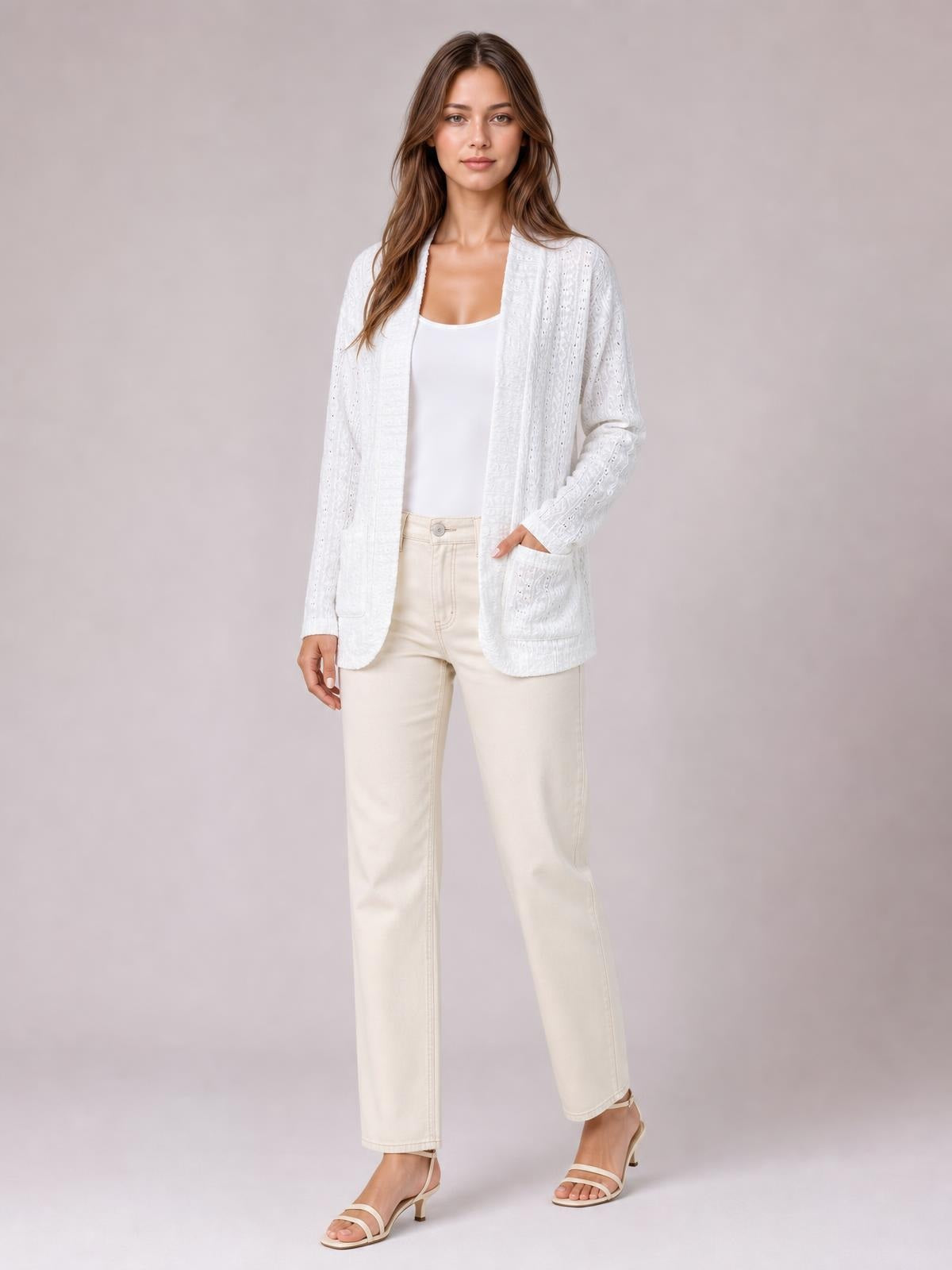 A model wears a white eyelet open-front cover-up with long sleeves, a relaxed silhouette, rounded hem, and large patch pockets, with one hand tucked inside.