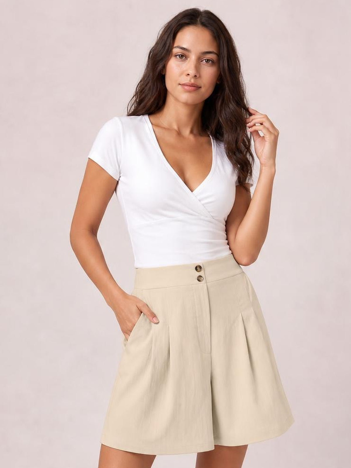 A woman modeling a white short-sleeve wrap-style top and beige high-waisted pleated shorts against a soft light pink studio background.