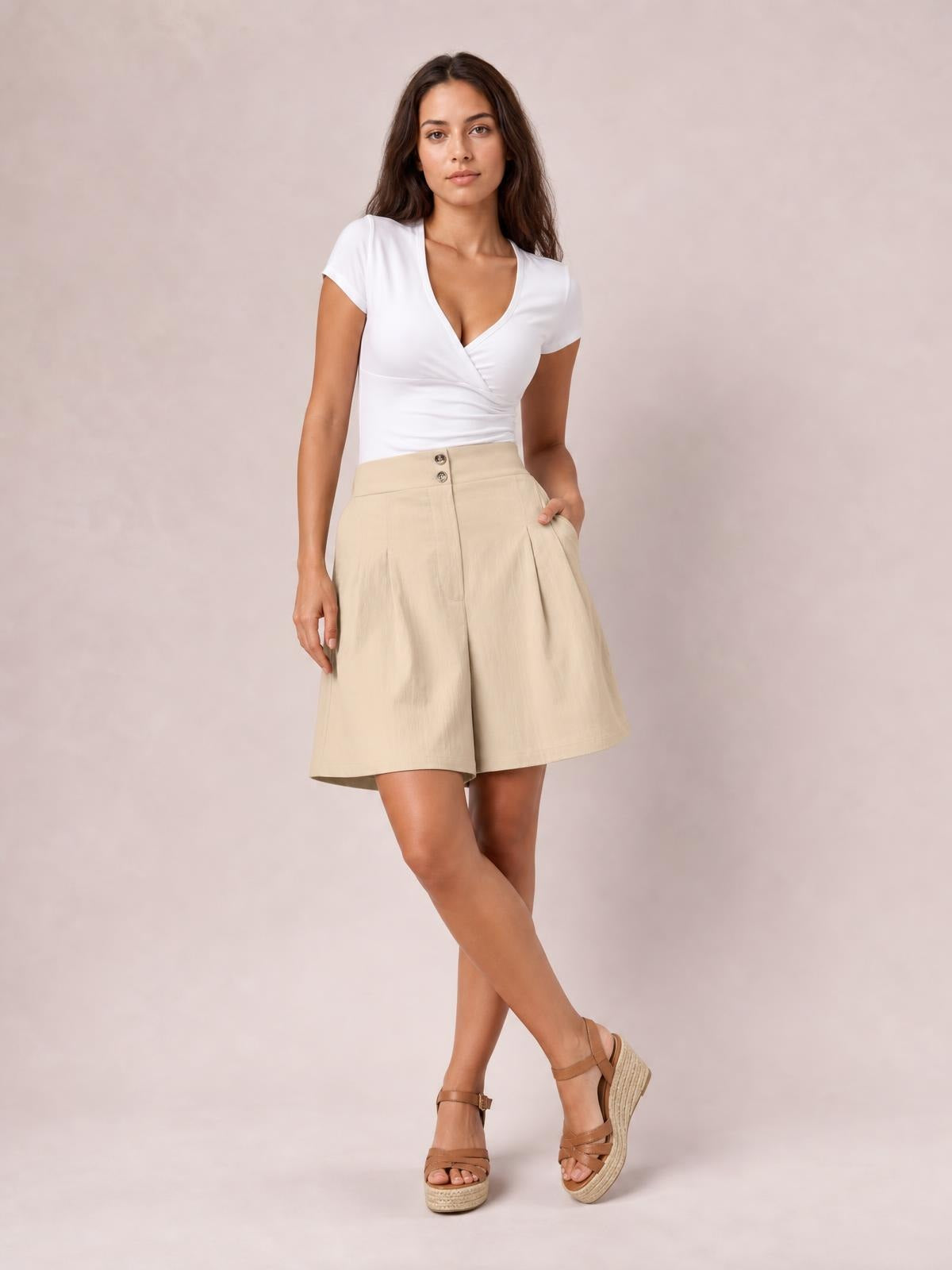 A model poses in beige pocketed shorts with a high-rise waistband, front buttons, soft pleats, and a relaxed wide-leg silhouette.