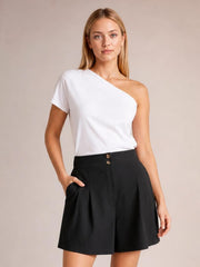 A woman modeling a white one-shoulder short-sleeve top paired with black pleated shorts against a solid beige studio background.