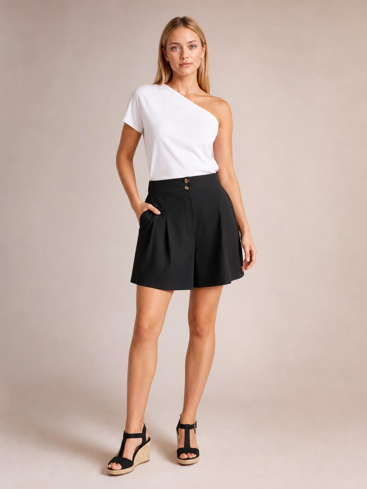 Model poses in black pocketed shorts with a high half-elastic waist, button closure, subtle ruching, and a relaxed wide-leg silhouette.