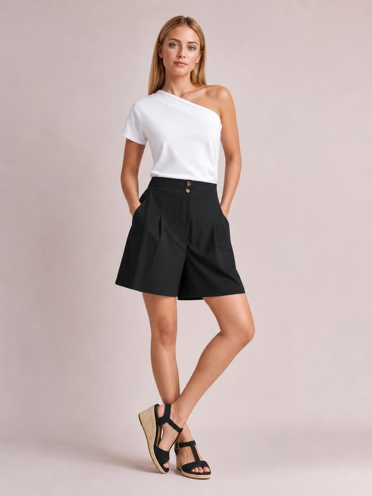 A model poses in black pocketed shorts with a high waist, front button closure, and wide-leg silhouette, crossing her legs beneath a white one-shoulder top.